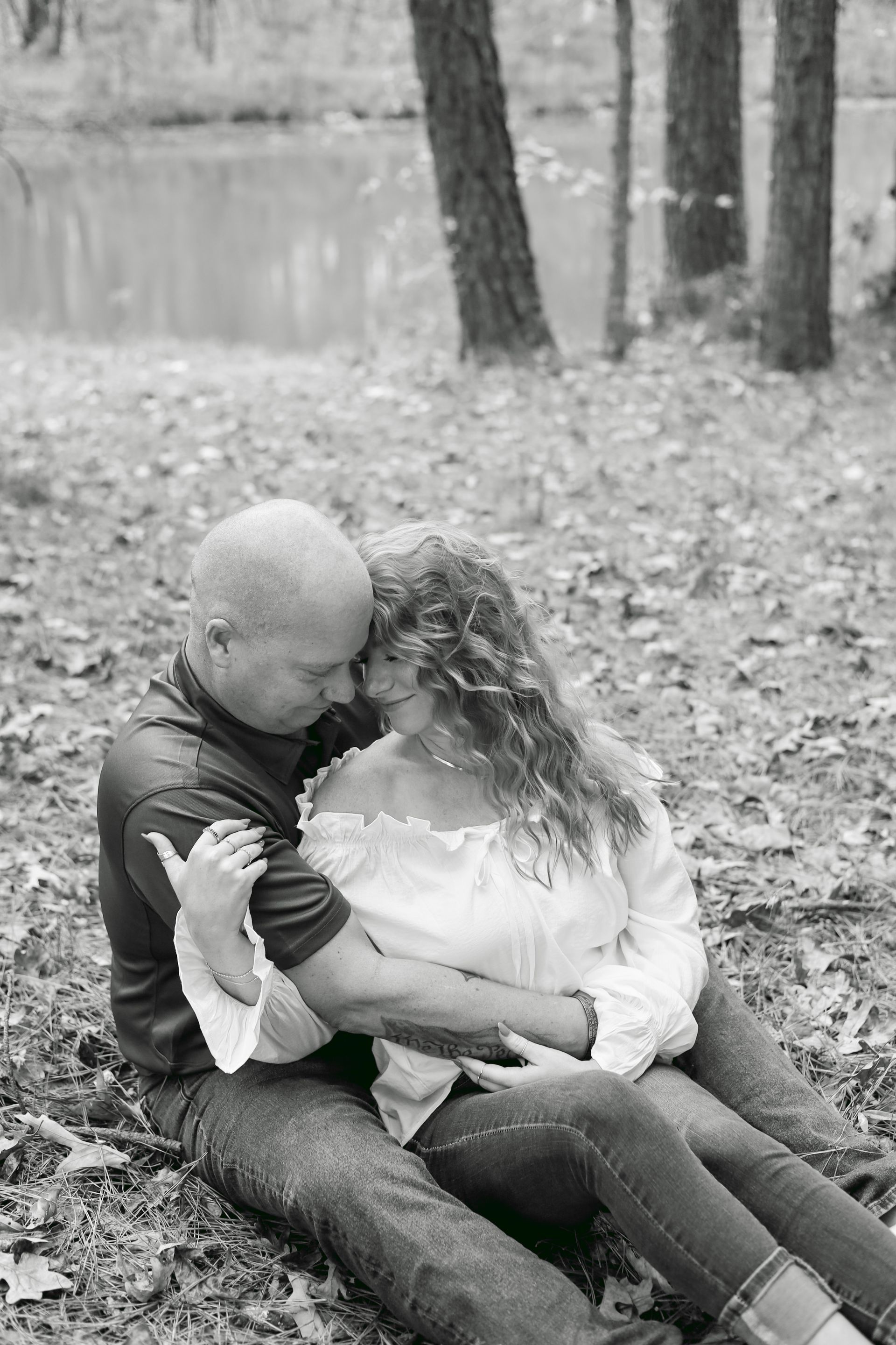 Couple embraces outdoors. Man in dark shirt, woman in white top, seated near trees and water.