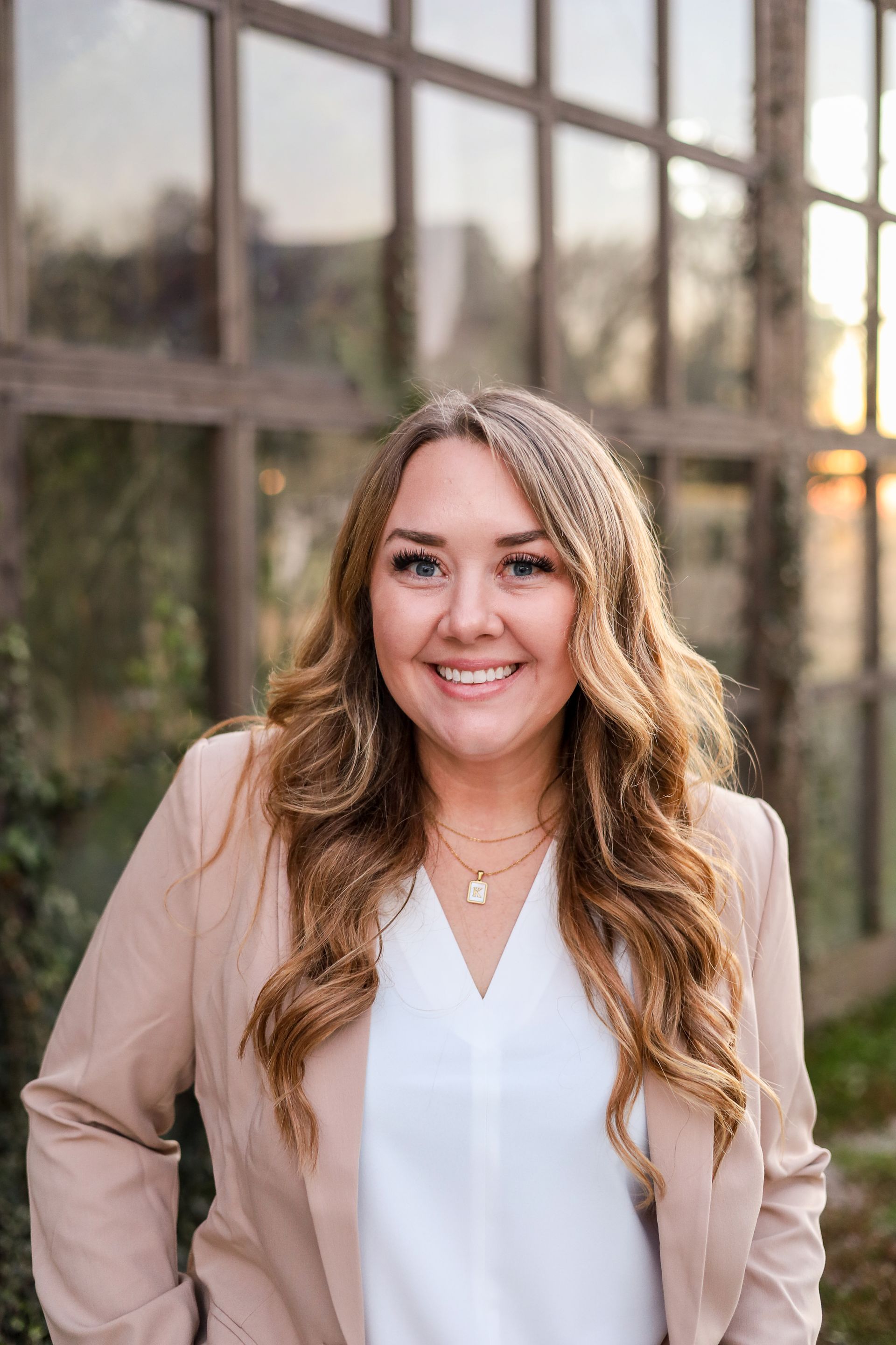 Woman in a tan blazer smiles, standing in front of a window.