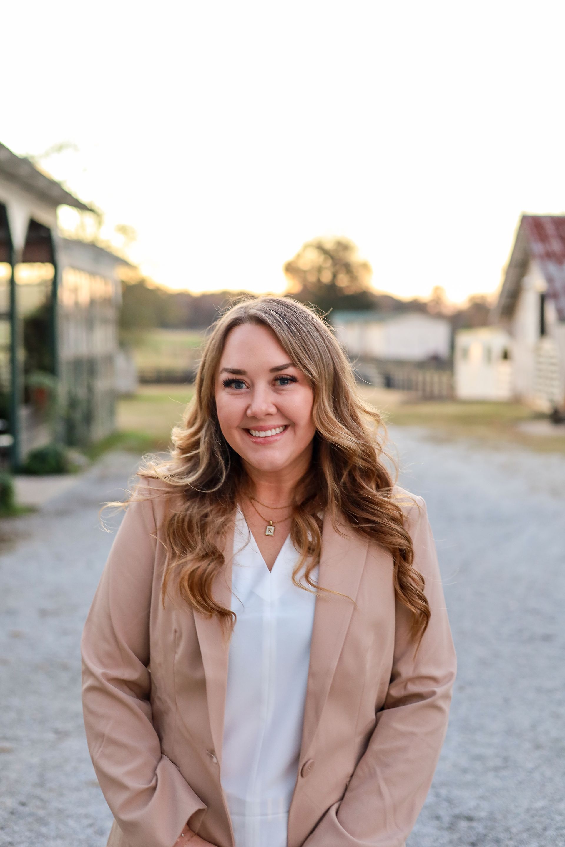 Woman smiling in a tan blazer and white shirt, standing outside with buildings in the background.