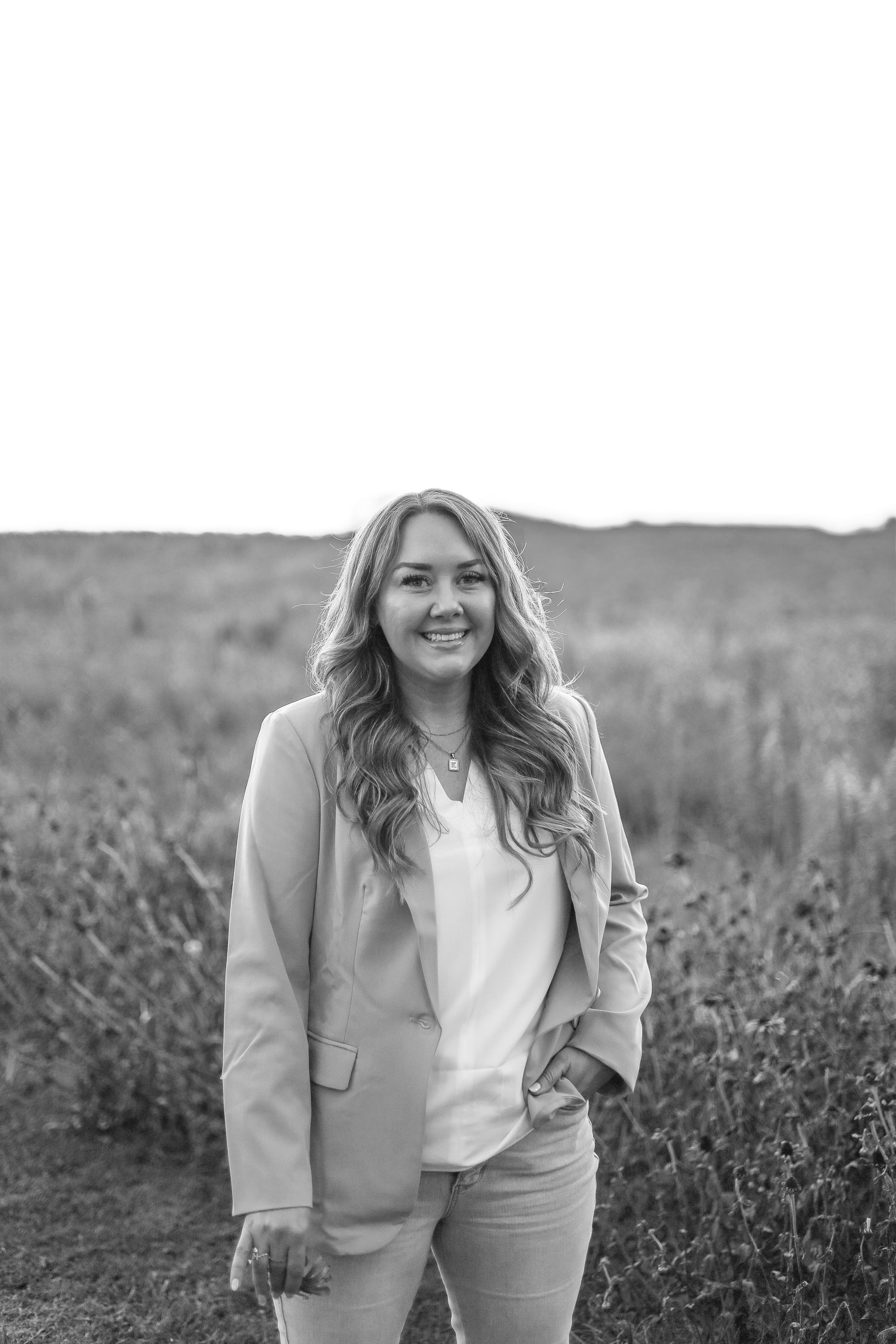 Woman in a blazer and jeans stands outdoors, smiling, hand in pocket, against a field backdrop.