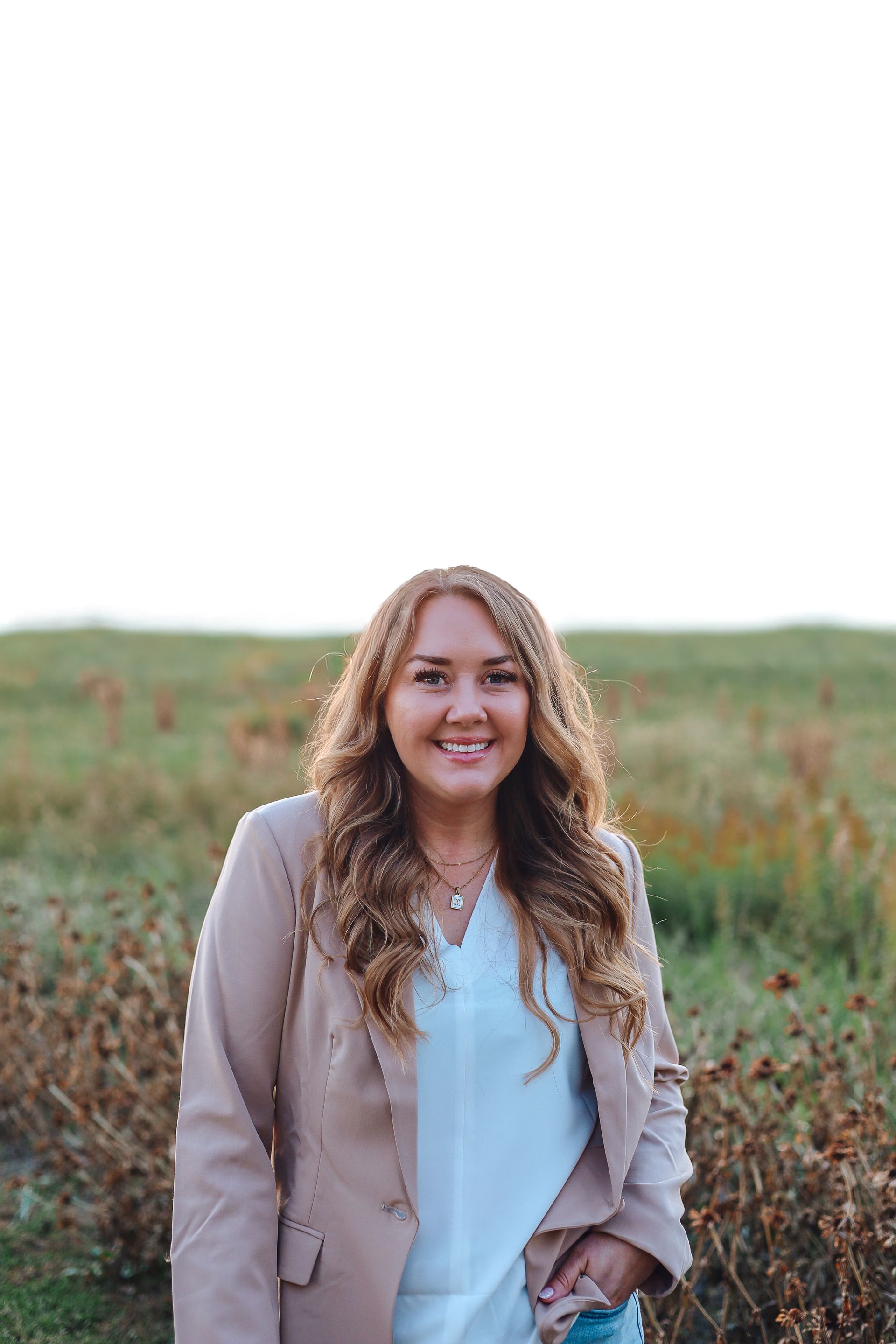 Woman in tan blazer and white shirt smiles outdoors in a field.