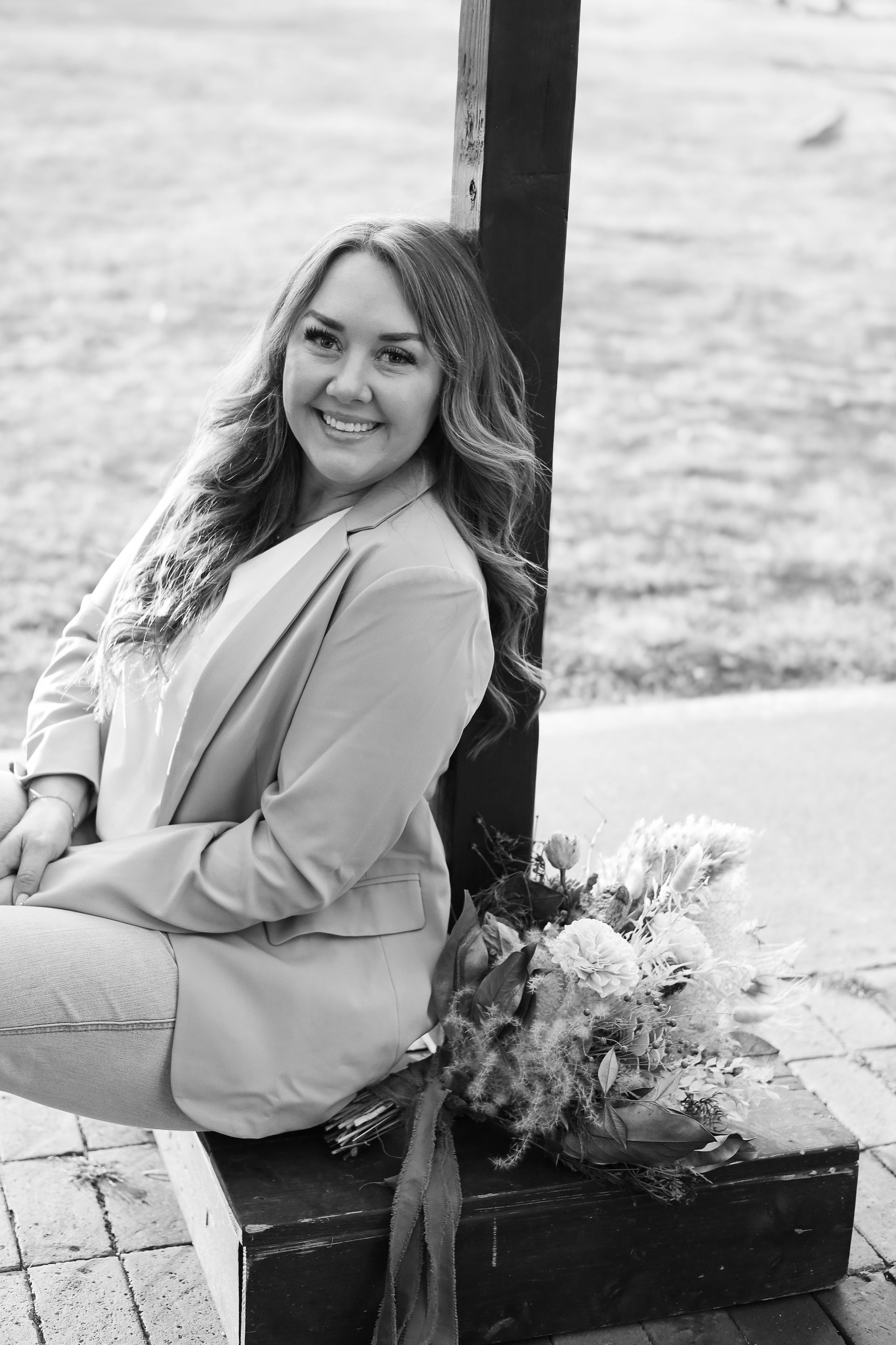 Woman in blazer leans against post, smiles, holding bouquet. Outdoor setting, black and white.