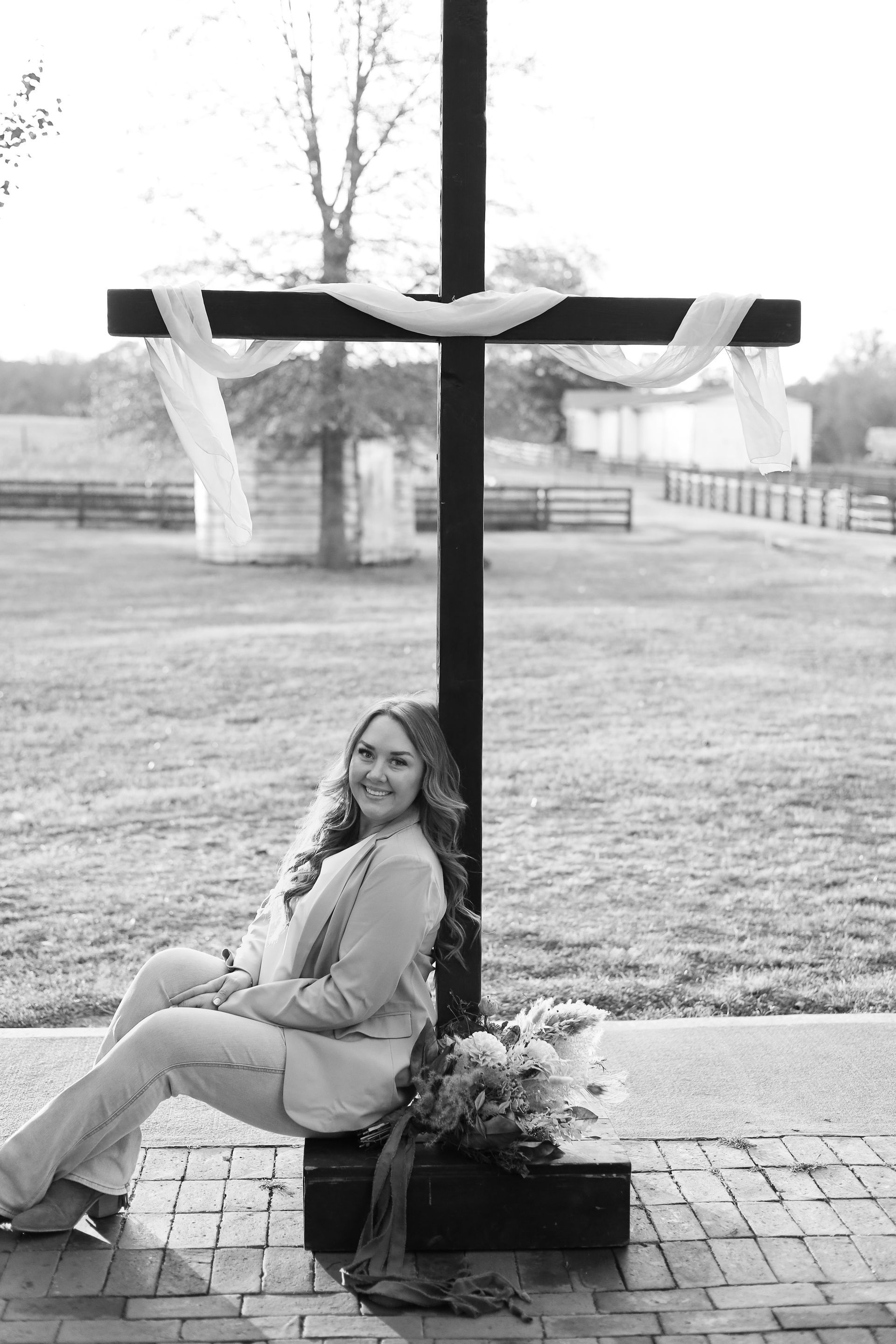 Woman seated beside a large wooden cross, smiling, bouquet of flowers at base, outdoor setting.