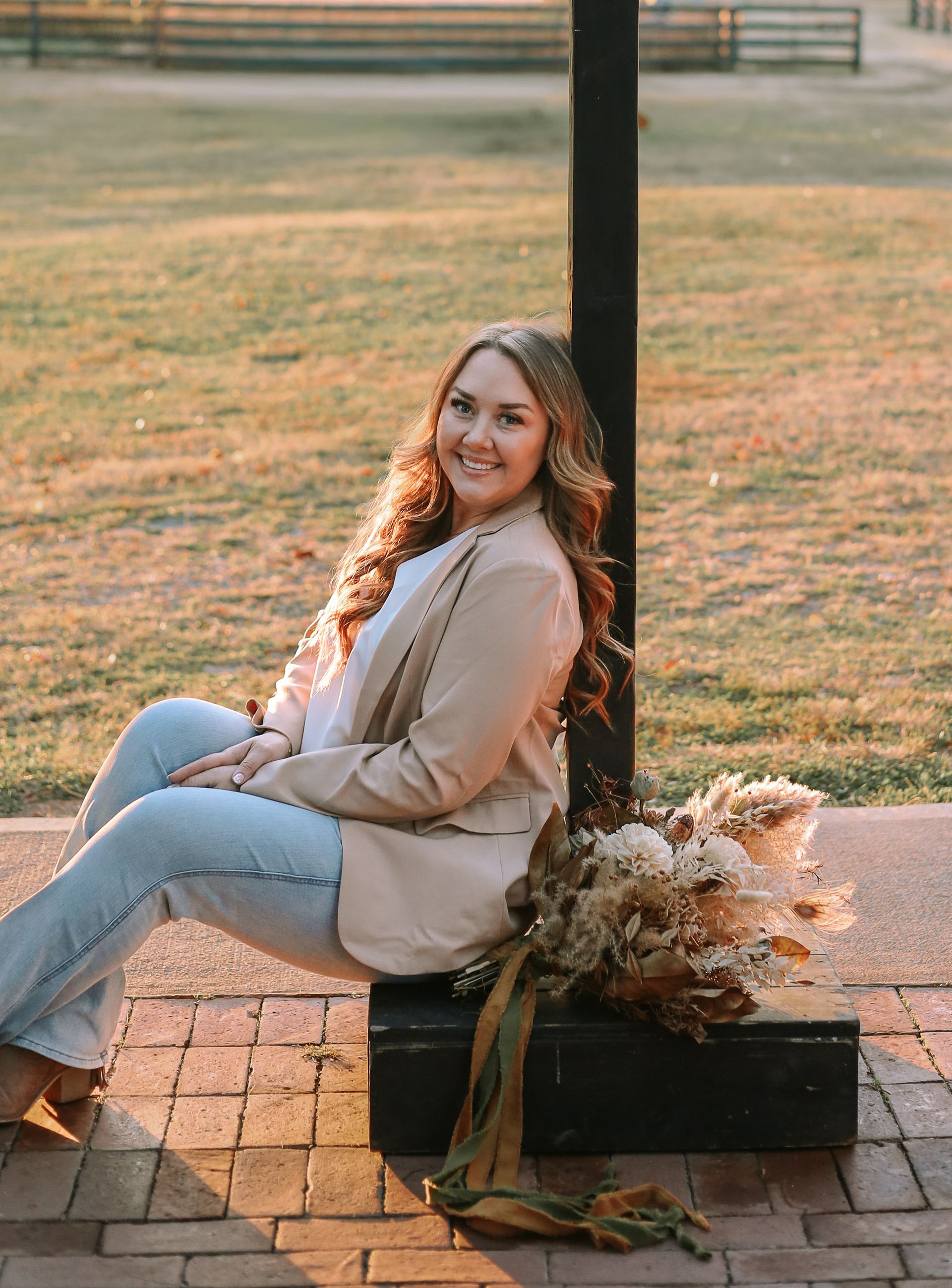 Woman in a tan blazer sits on a brick surface, leaning against a black pole with flowers.