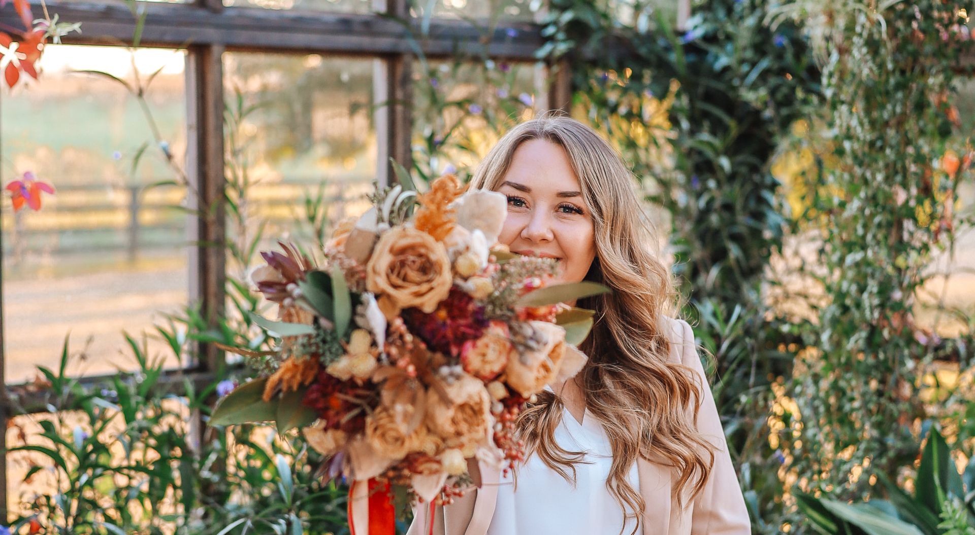 Woman with curly hair holds a fall bouquet in front of a window, smiling.