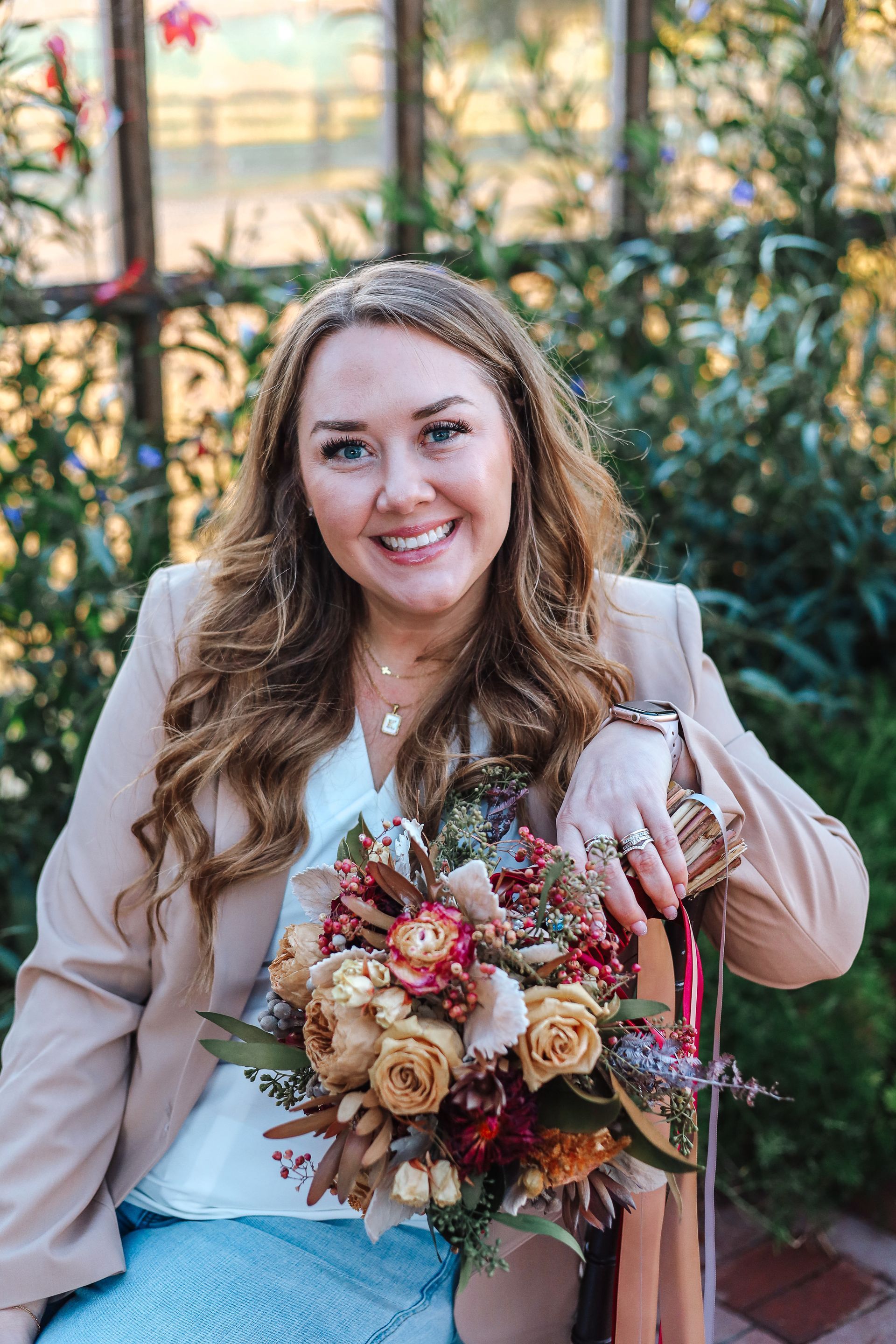 Woman smiling, holding a bouquet, wearing a blazer, seated outside near greenery.