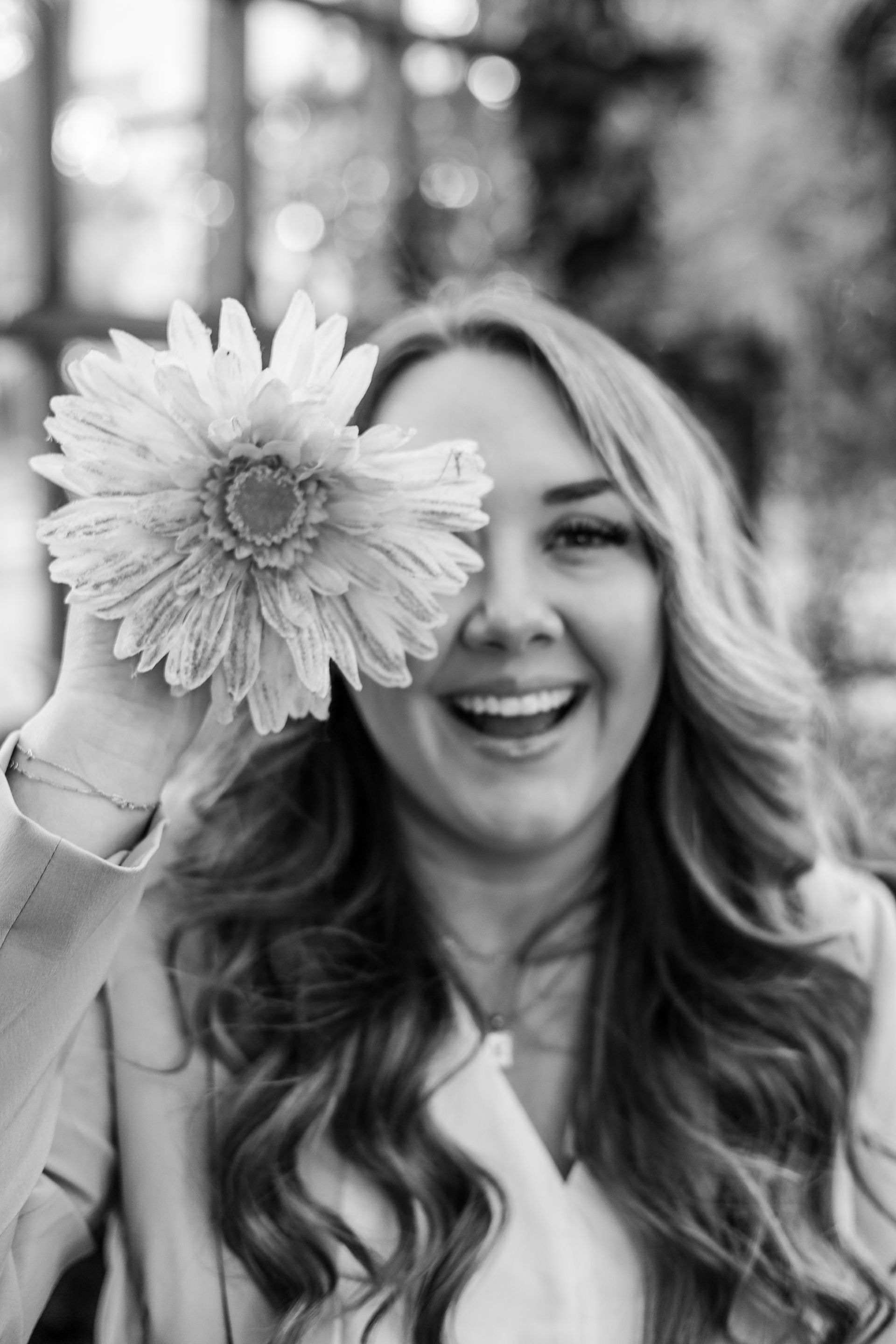 Woman holding a large daisy over one eye, smiling broadly, outdoors.