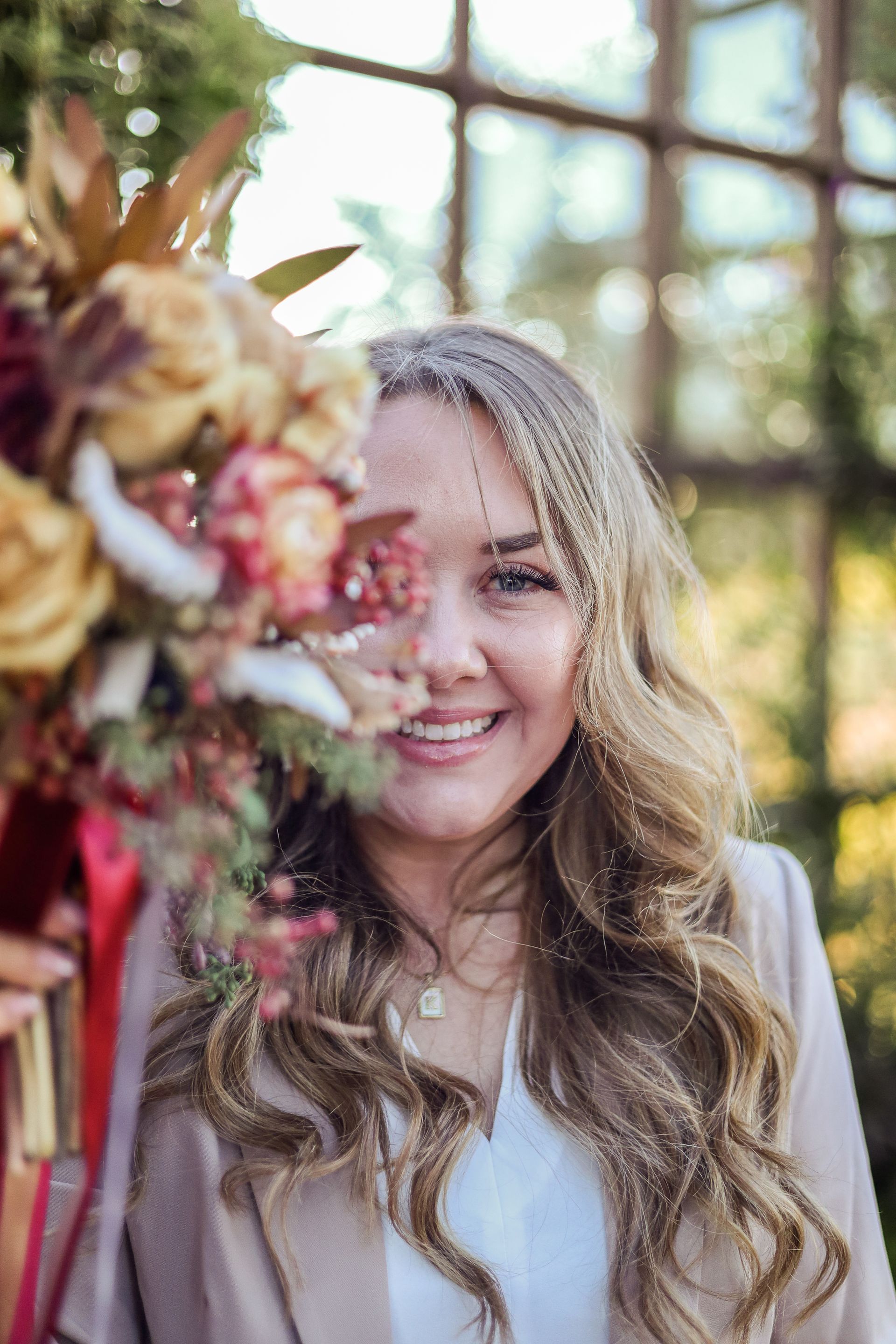 Woman smiling, holding bouquet, in front of a window. Warm colors and sunlight.