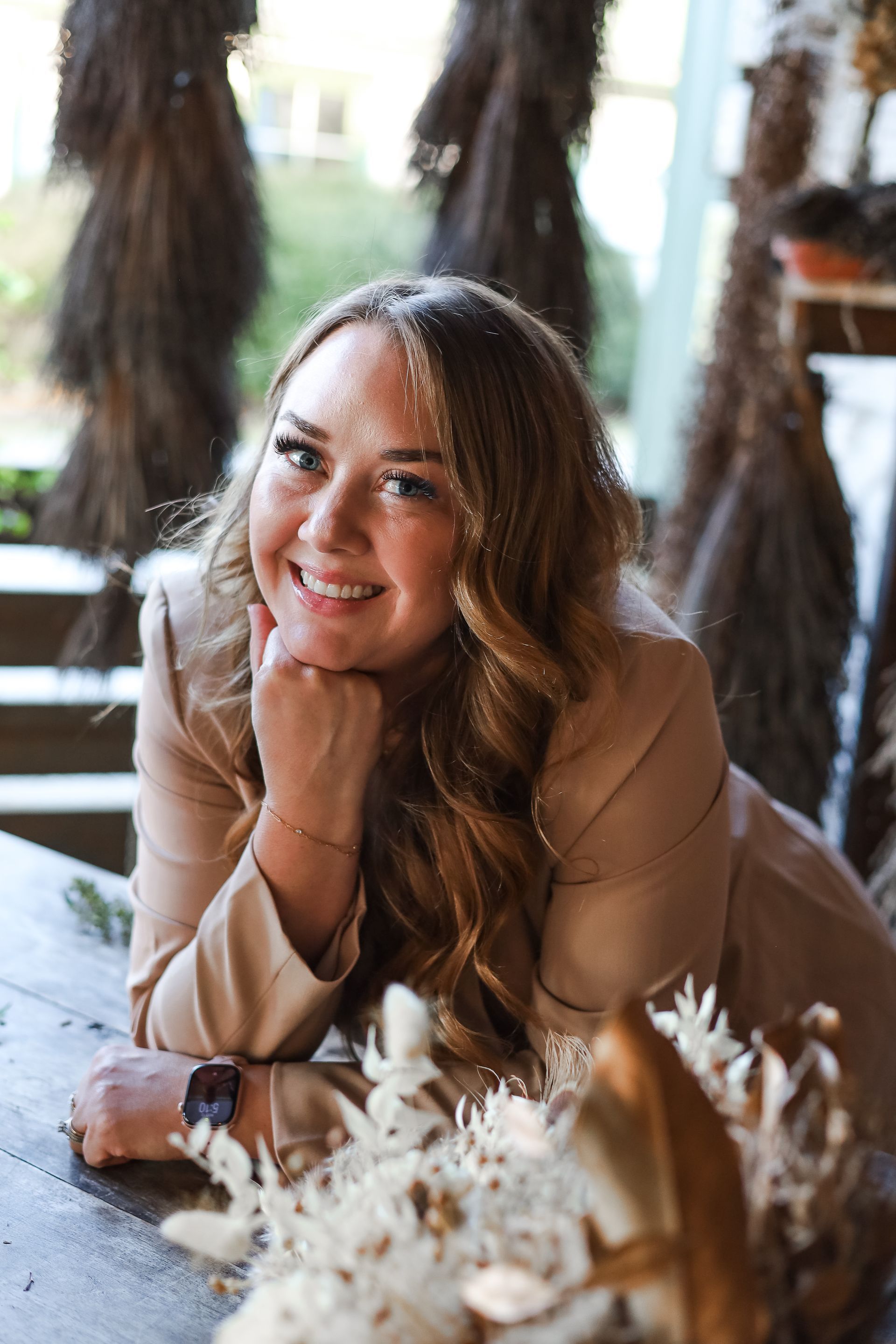 Woman in a tan blazer smiles, resting her chin on her hand, leaning on a wooden table.