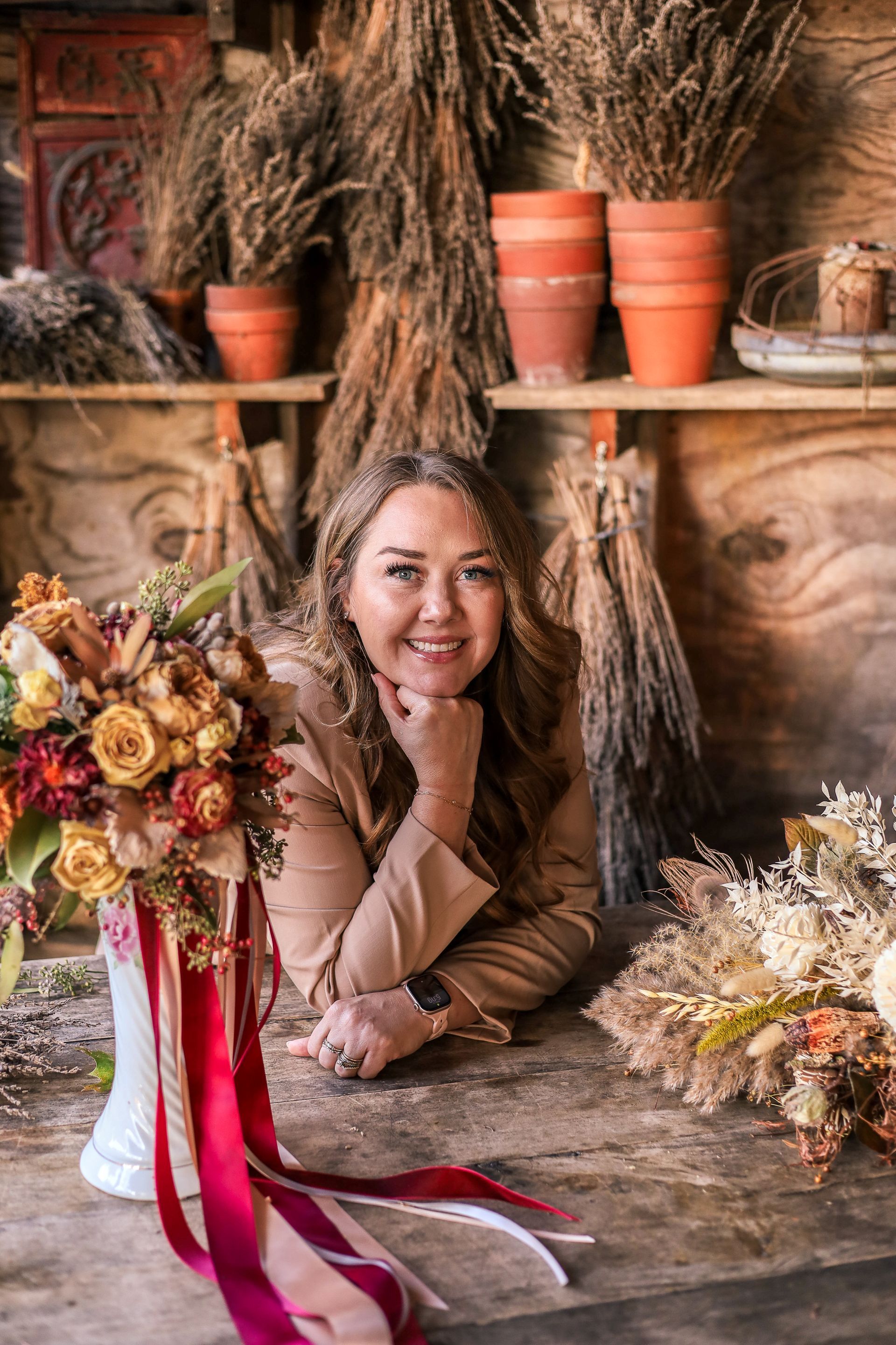 Woman smiles, leaning on wooden table with floral arrangements. Beige jacket, pottery, dried plants.