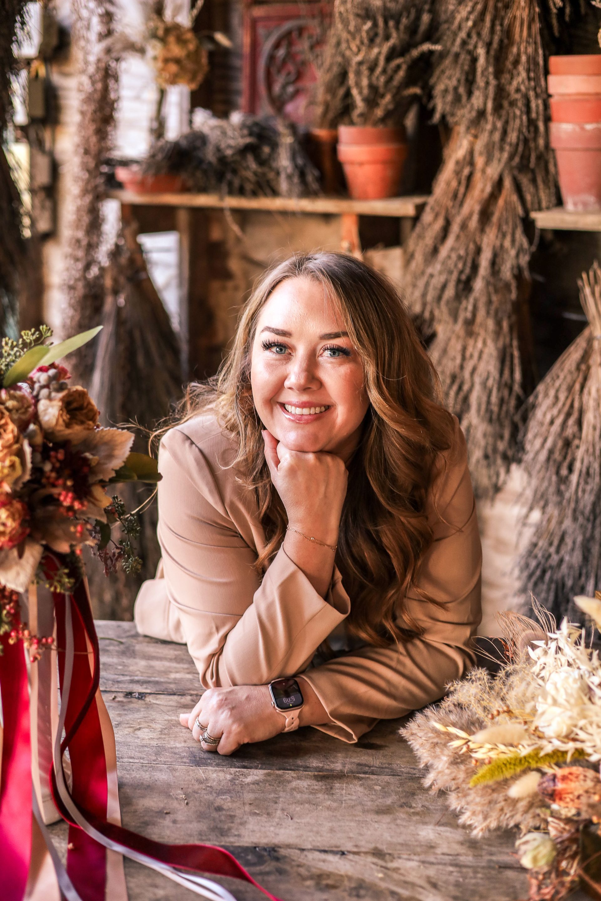 Woman in tan blazer smiles, resting her chin on hand, in a rustic, floral setting.