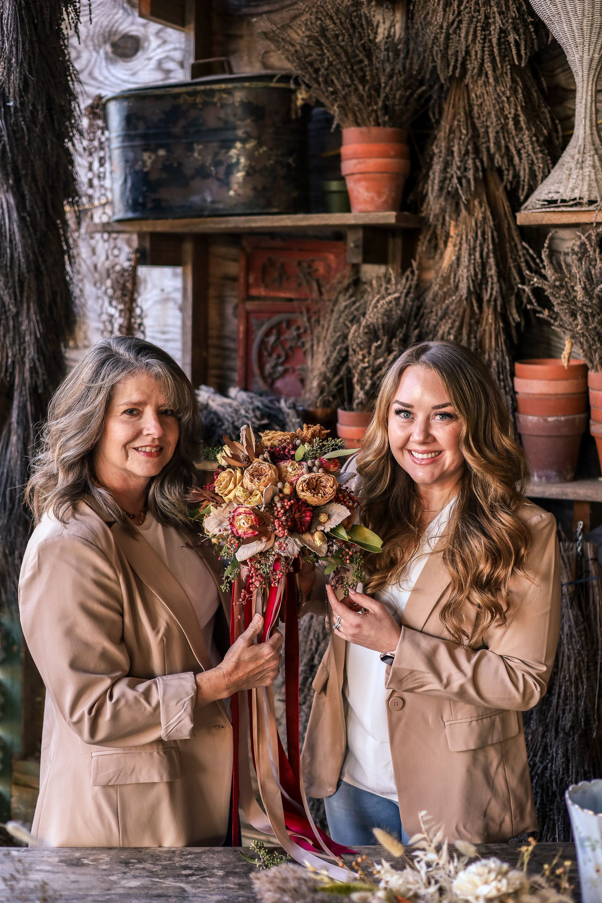 Two women in tan blazers hold a bouquet of flowers in a rustic shop.