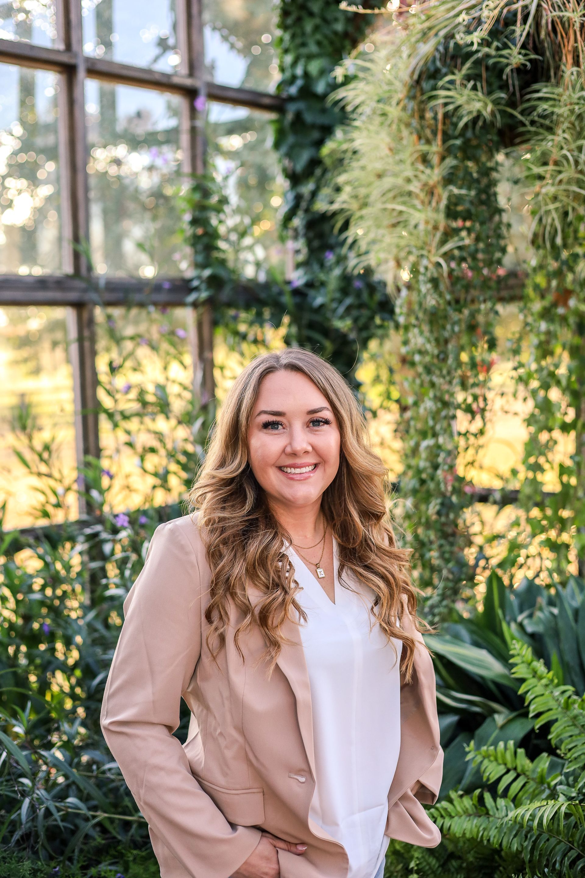 Woman in a light blazer smiles in front of a window and greenery.