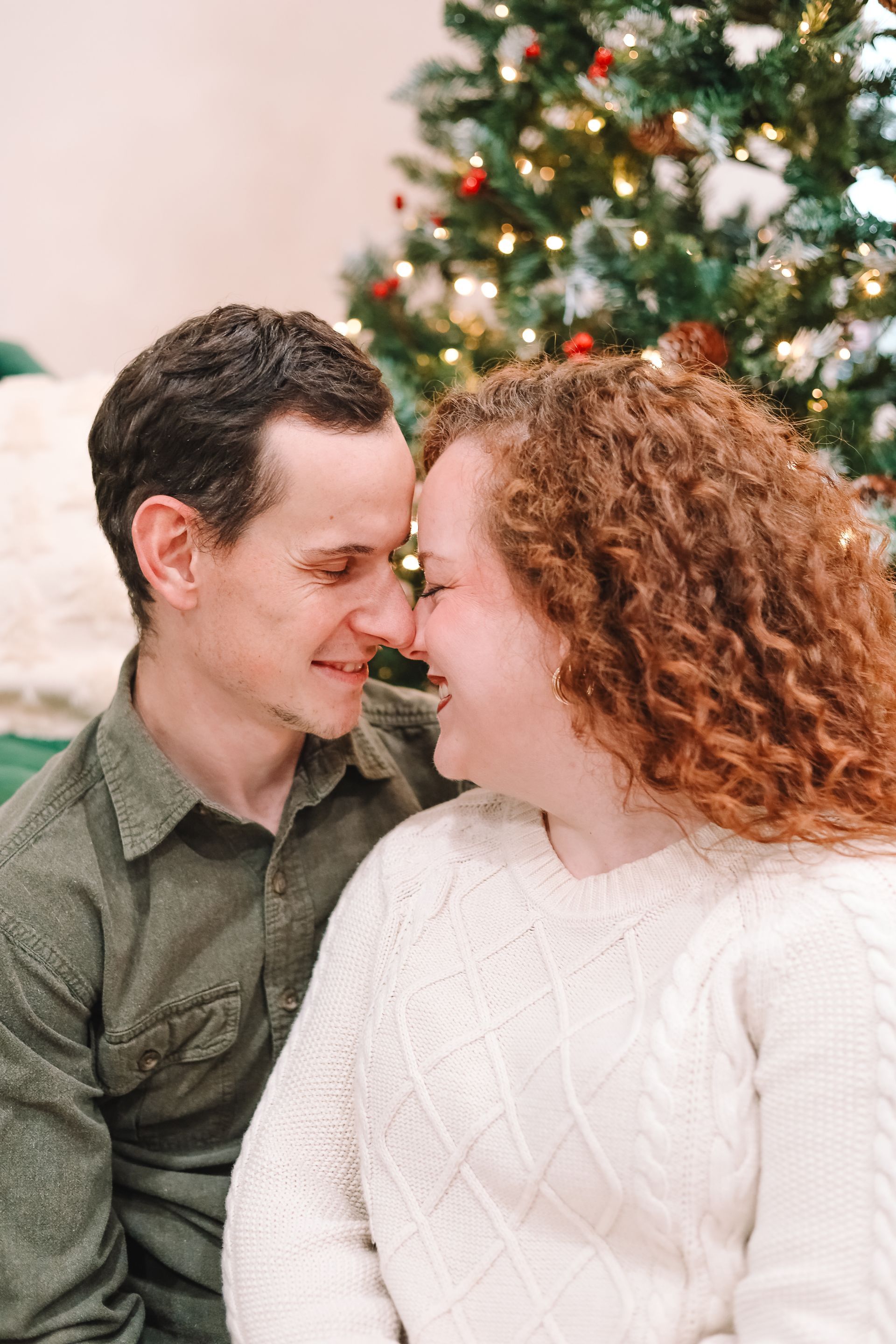 Couple smiling, noses touching, by a Christmas tree. Man in green shirt, woman in white sweater.