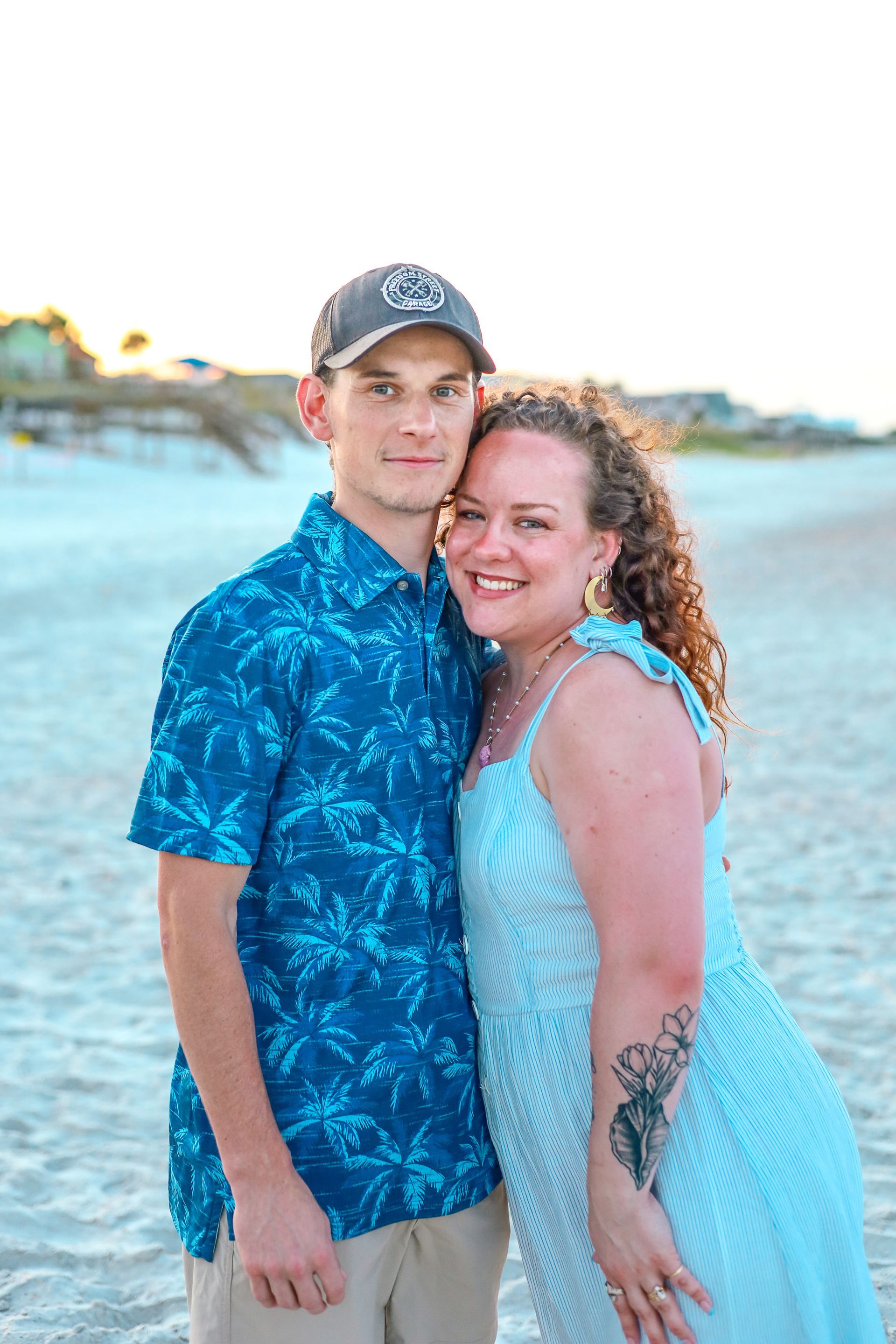 Couple embraces on a beach, woman in white dress, man in blue shirt, sun setting.