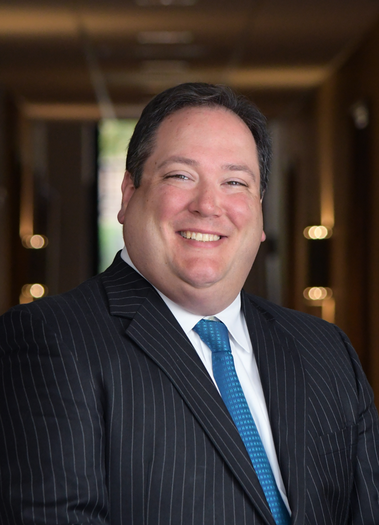 A smiling person wearing a navy blue pinstripe suit, white shirt, and blue tie, standing in a softly lit office hallway.