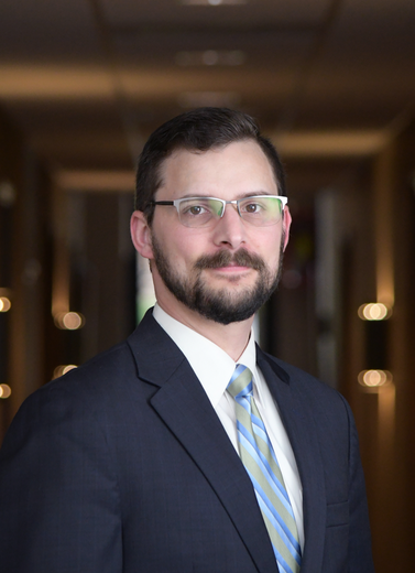A person with a beard and glasses wearing a dark suit and striped tie, posed in front of a softly lit corridor.