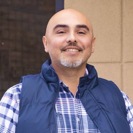 A bald man with a beard is wearing a plaid shirt and a blue vest.