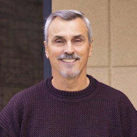 A man with a beard and mustache is wearing a purple sweater and smiling.