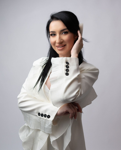 Woman in white blazer smiles, hand in hair, against a light background.