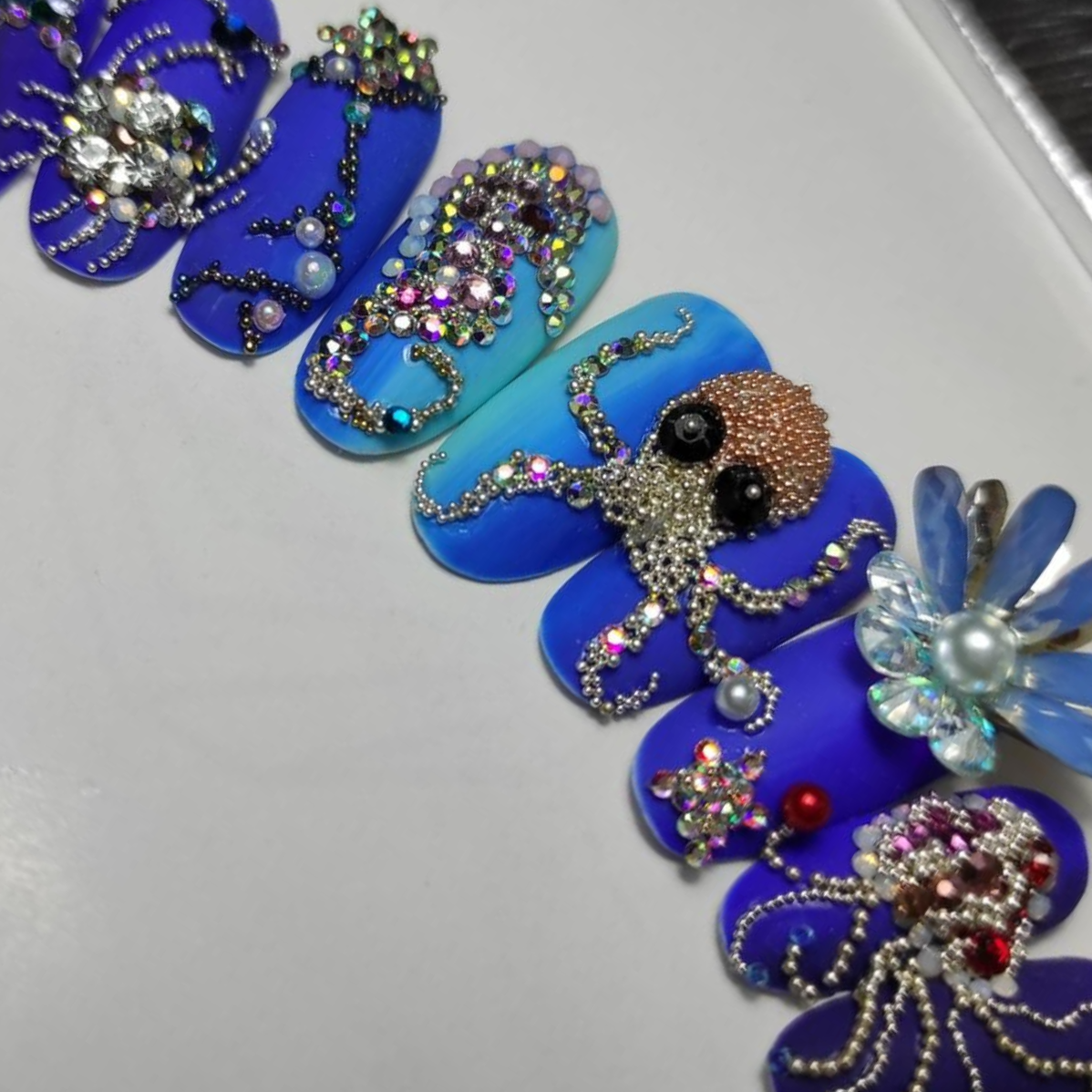 Elaborate, bejeweled ocean-themed nail art with shades of blue, featuring octopuses, pearls, and sparkling accents.