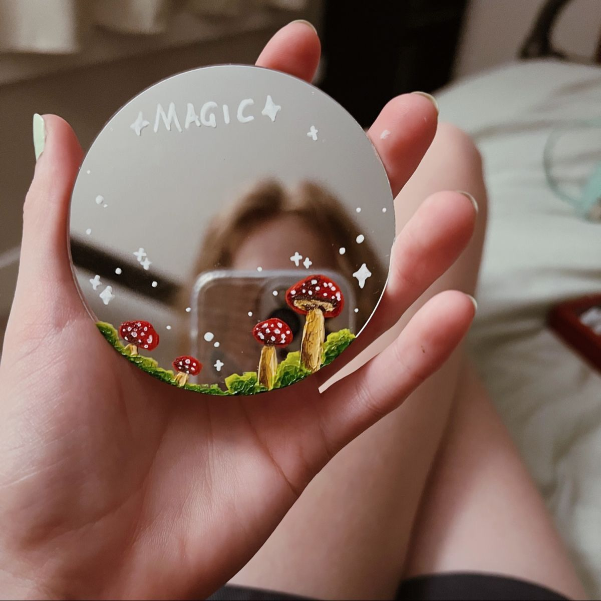Hand holding a round mirror painted with mushrooms and the word