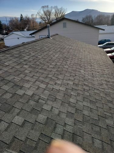 Gray asphalt shingle roof on a house, with chimney and surrounding buildings visible under a blue sky.