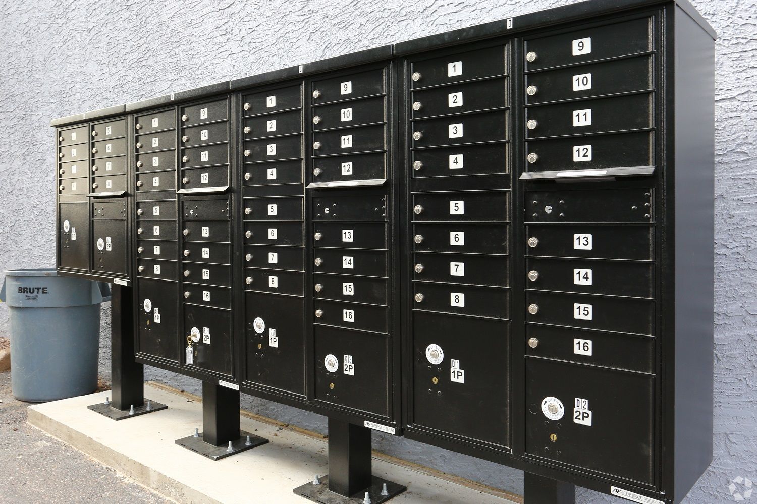 Black mailboxes with numbered slots, mounted on black posts, next to a trash can.