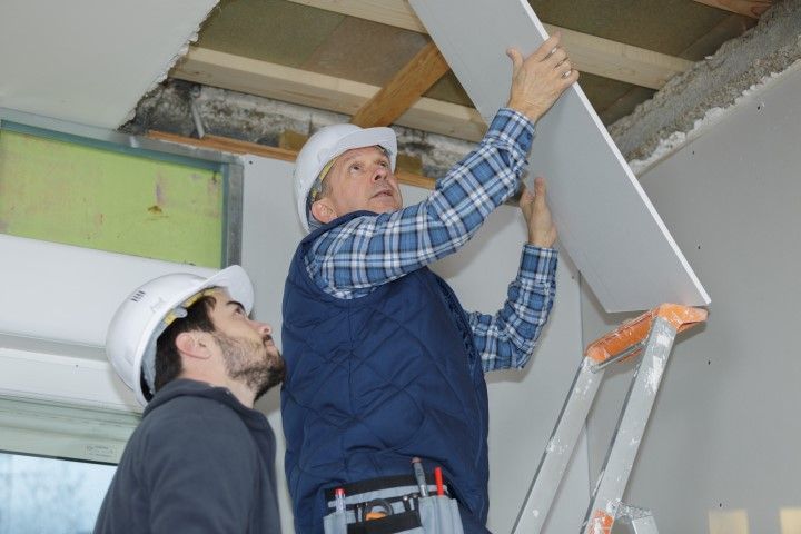 Two construction workers install a drywall panel on a ceiling; one holds it in place while the other observes.