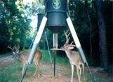 Two deer are standing next to a deer feeder in the woods.