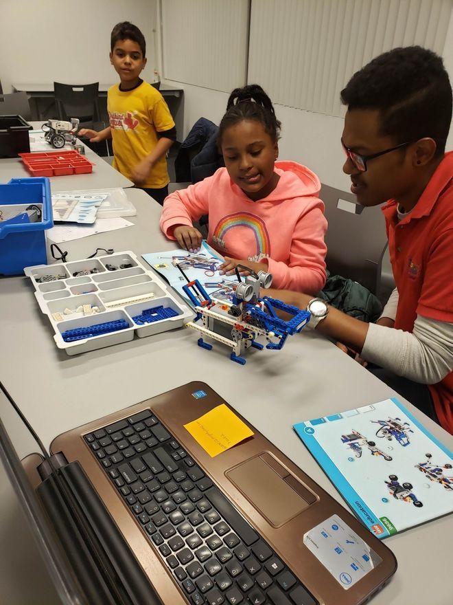 A mentor and two children build a robot together at a desk with instruction manuals, LEGO pieces, and a laptop.