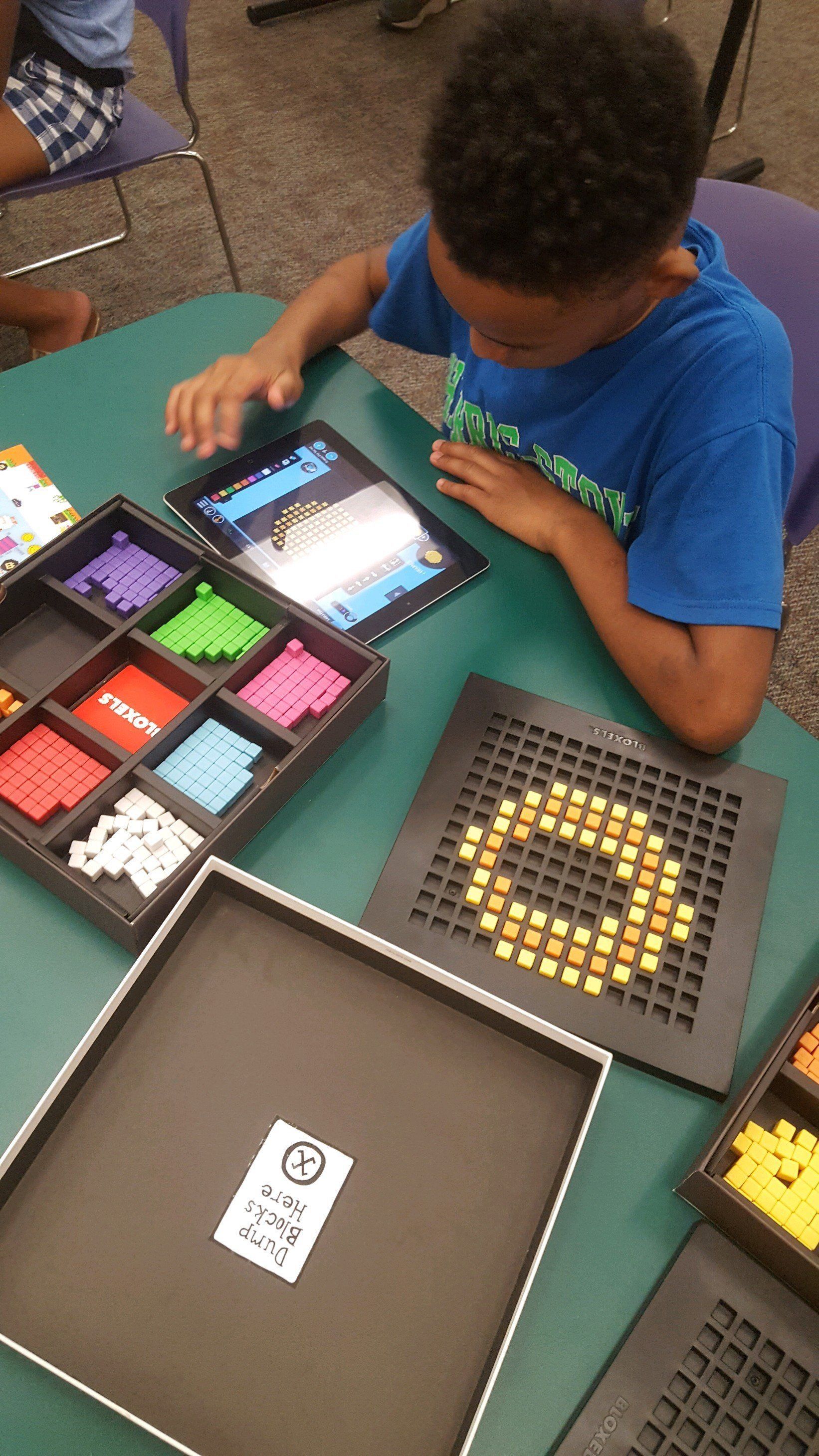 A student uses a tablet to arrange colorful plastic tiles on a grid board during an educational activity.