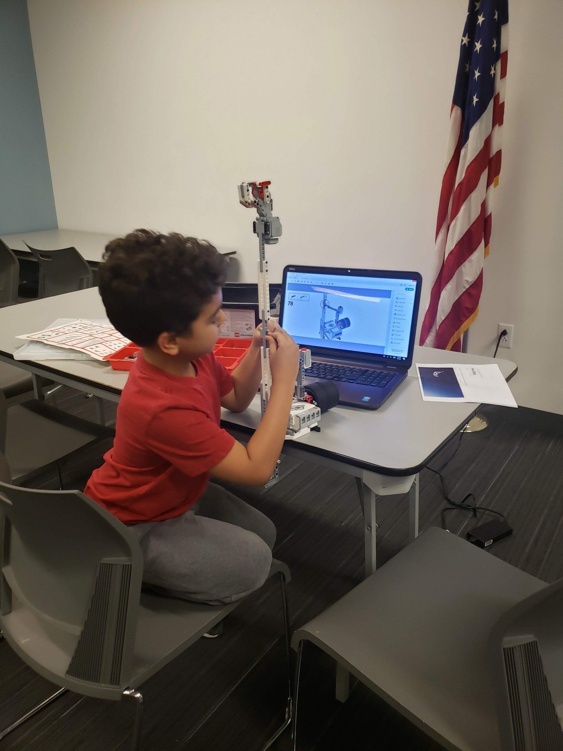A child in a red shirt works with a robotics kit next to a laptop on a table with a US flag in the background.