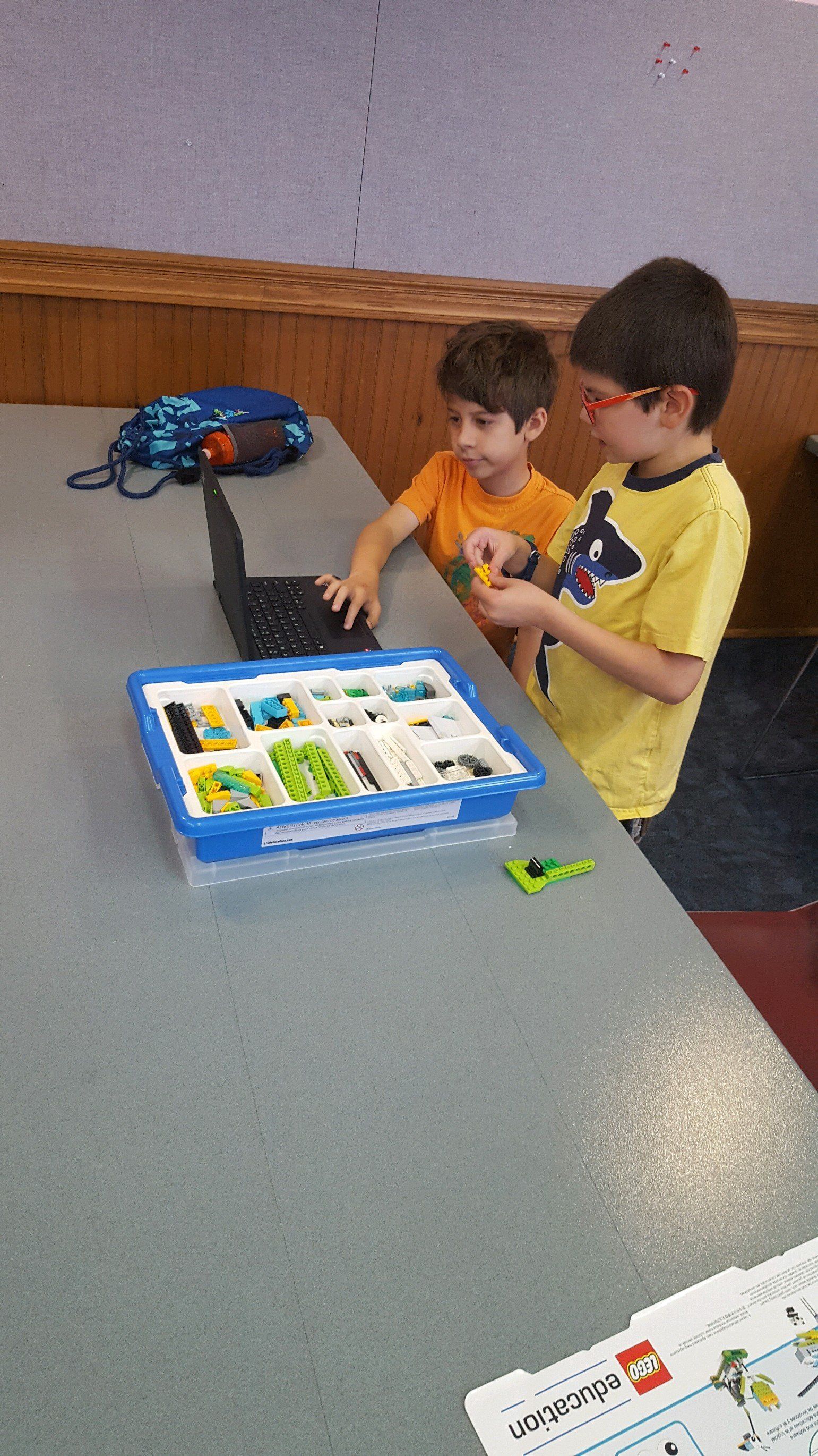 Two children work together at a table on a project using a laptop and a divided tray of colorful building bricks.