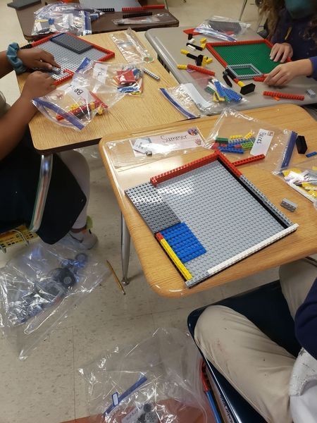 Students work at desks with LEGO baseplates, assorted colorful bricks, and plastic bags of building supplies.