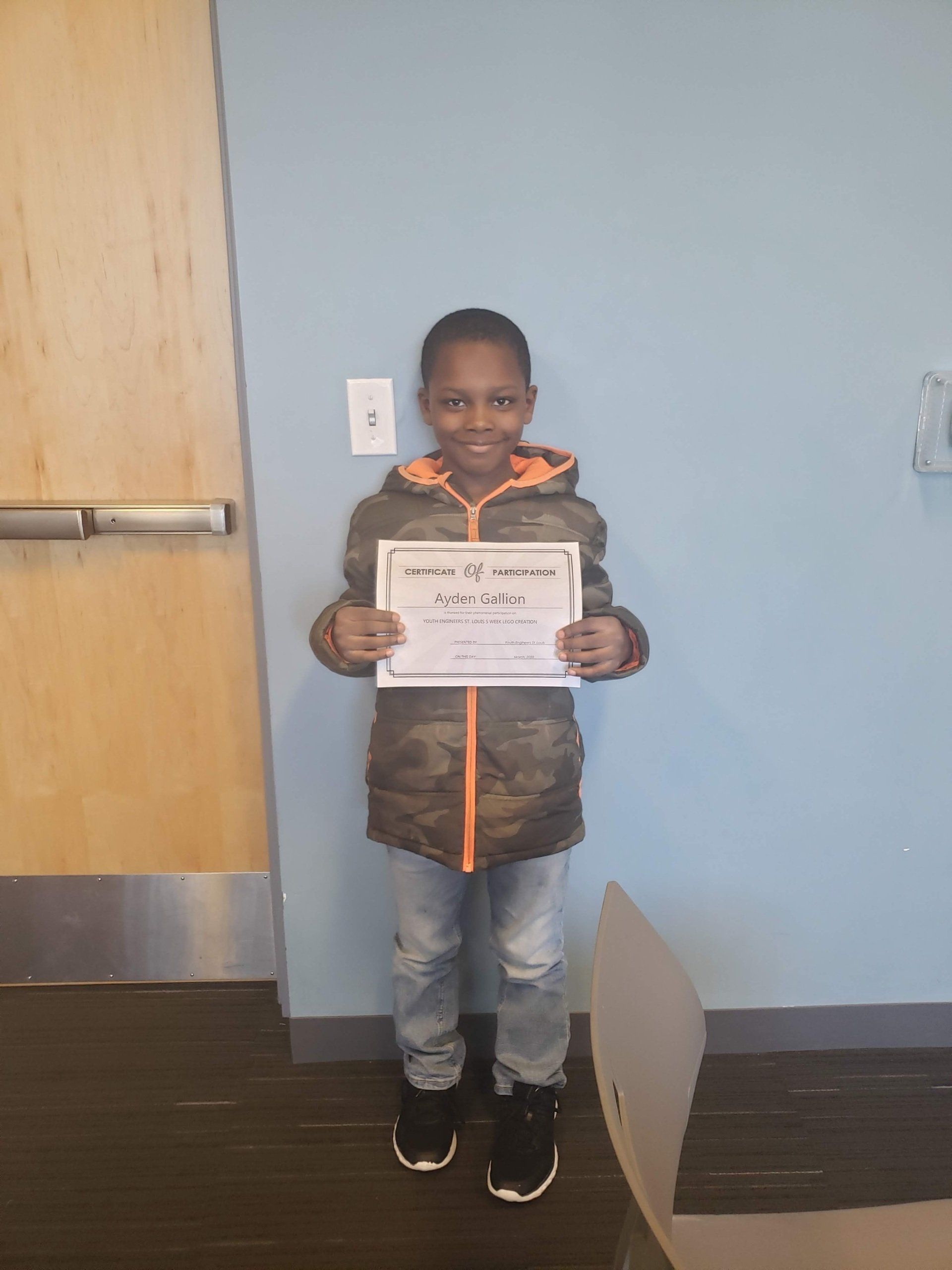 A student wearing a camouflage jacket stands against a blue wall, smiling while holding a certificate.