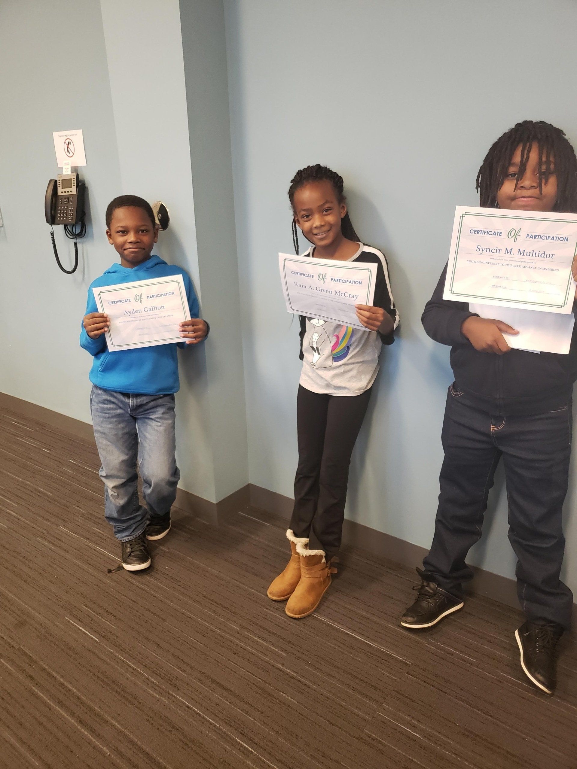 Three individuals stand against a light blue wall, each holding a handwritten sign.