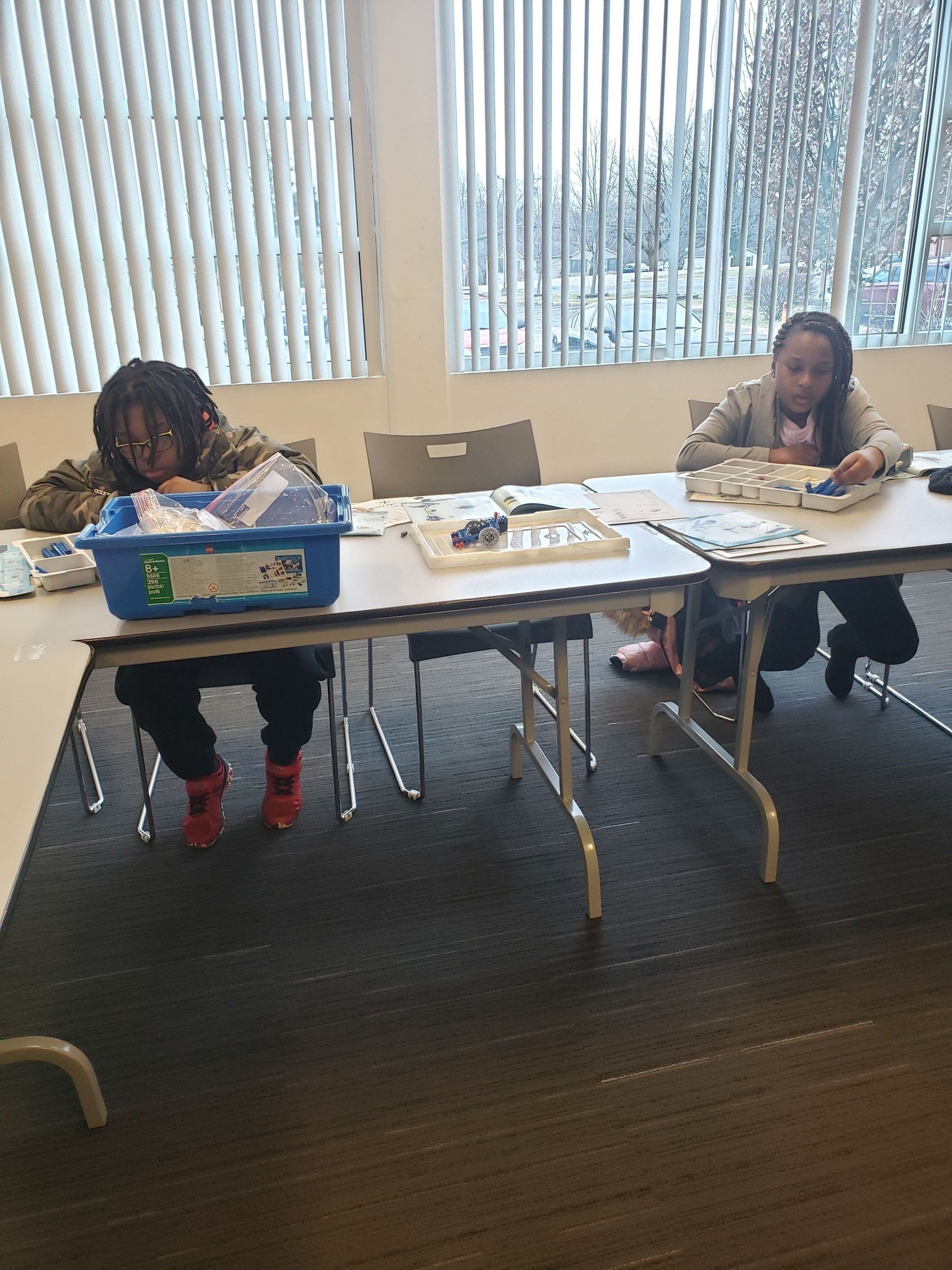 Two people sit at tables in a brightly lit room, focused on organizing small items into containers and trays.