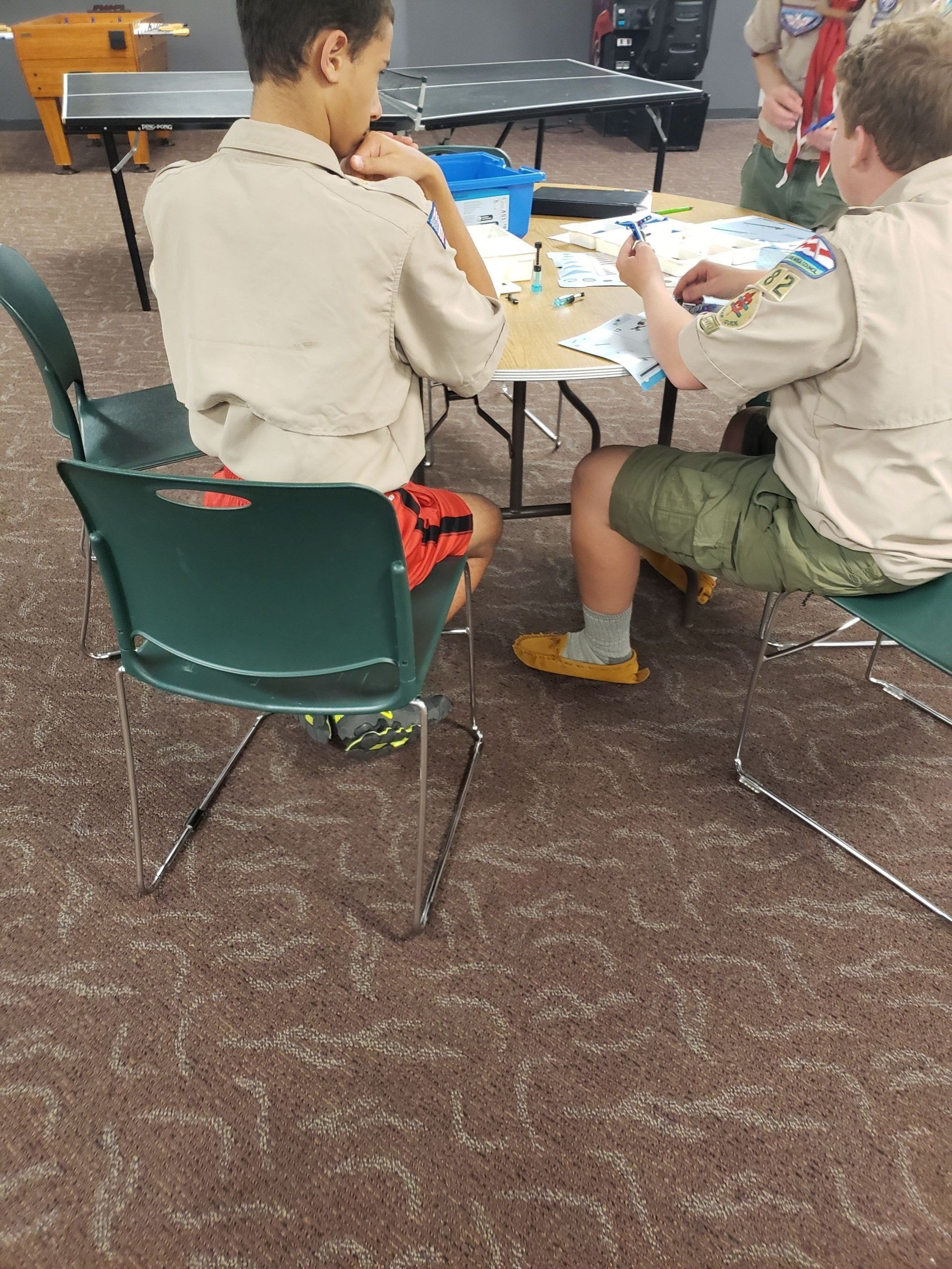Two scouts in uniform sit at a table in a room, working on an activity with papers and pens.