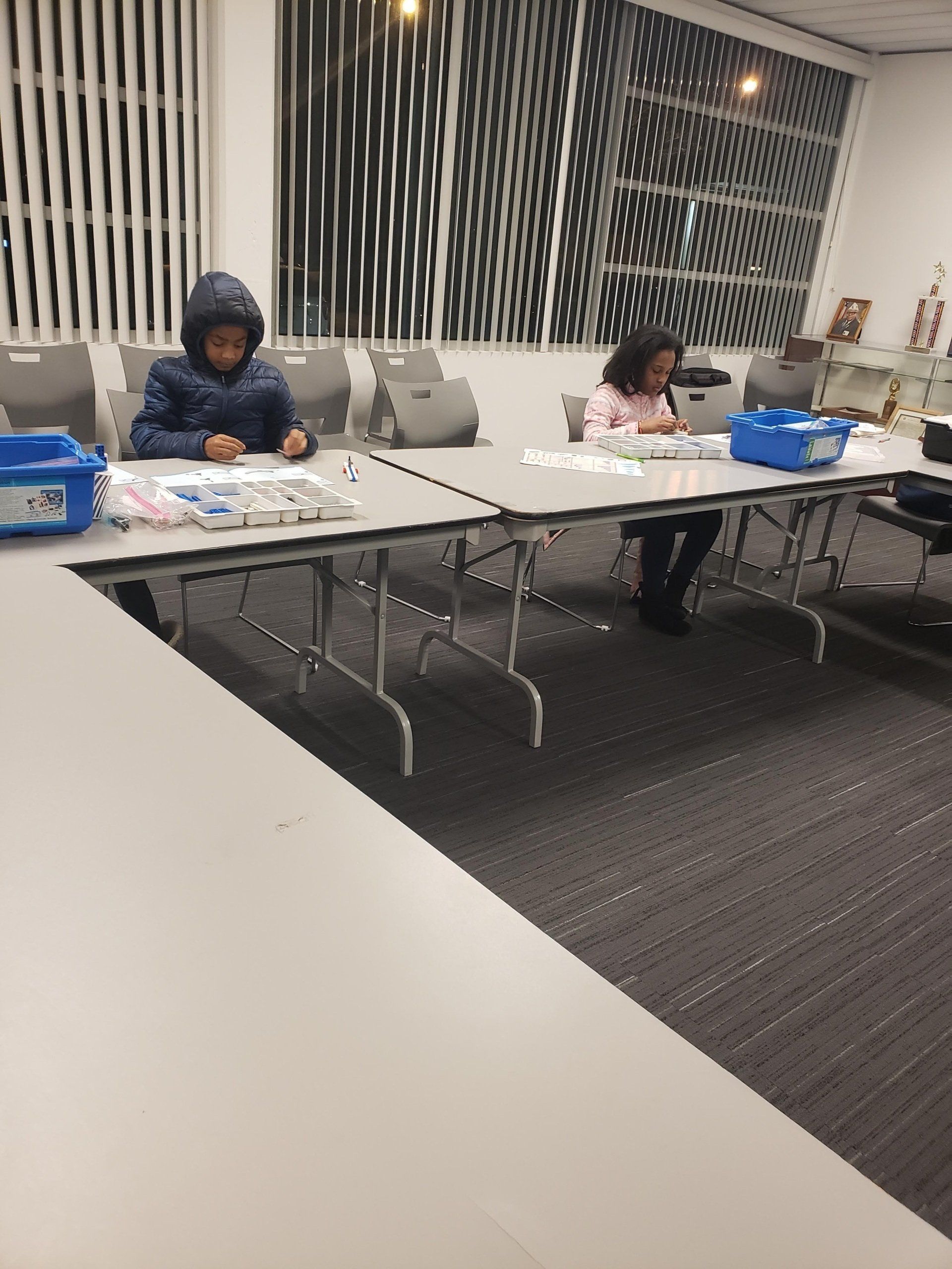 Two people sit at tables in a room, focused on small, organized items arranged on their workspaces.