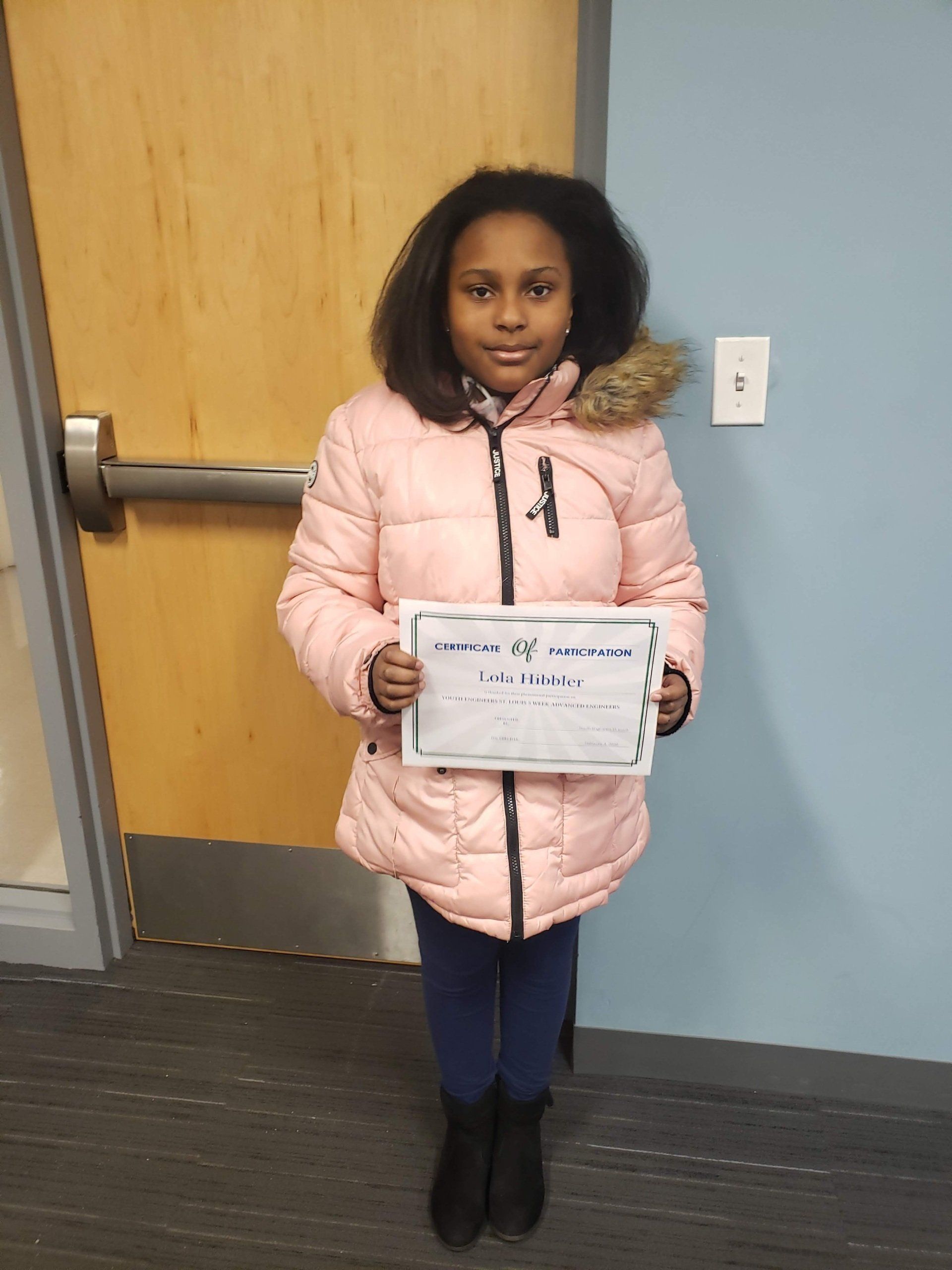 A person wearing a light pink puffer jacket holds a piece of paper in front of a wooden door and light blue wall.