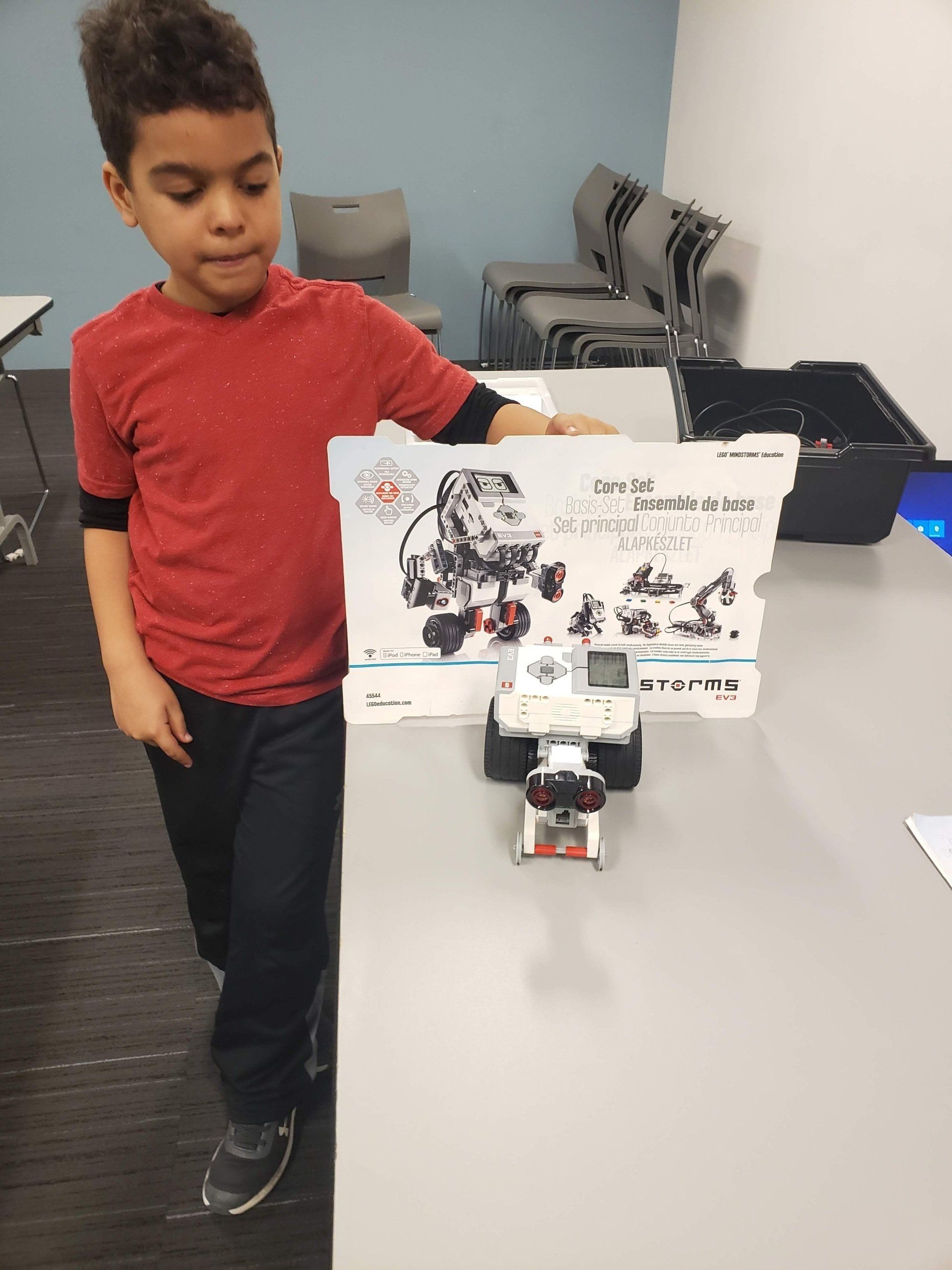 A child in a red shirt shows a small, white robotic vehicle and a matching instruction card on a table in a room.