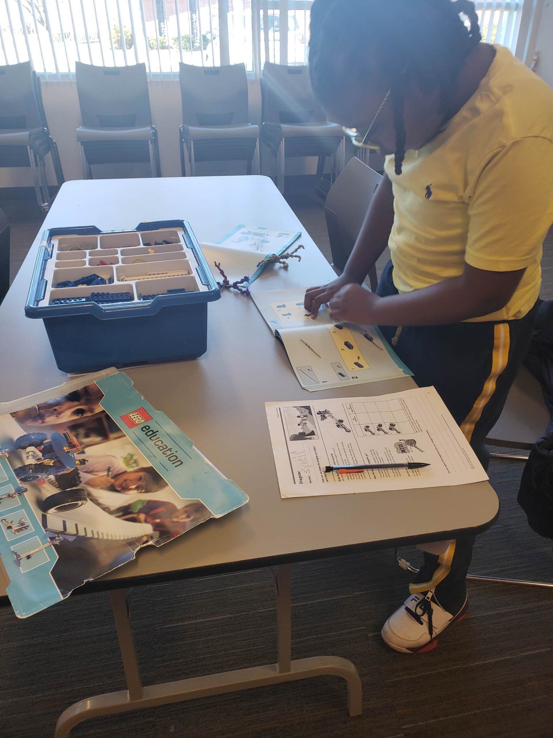 A person in a yellow shirt assembles a Lego set at a table with an instruction manual and a storage tray.