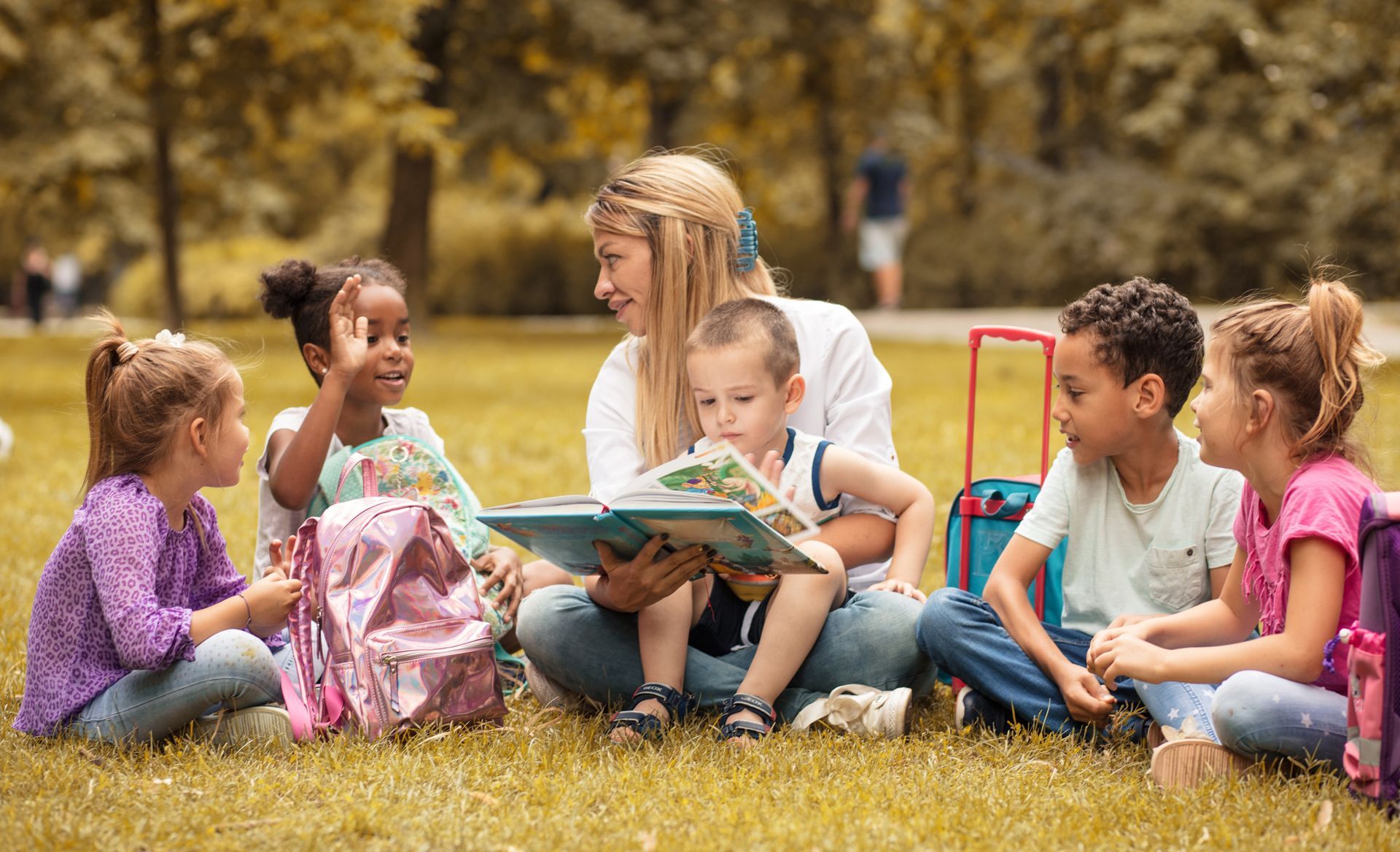 A teacher reads a book to four children sitting in a circle on the grass in a park.