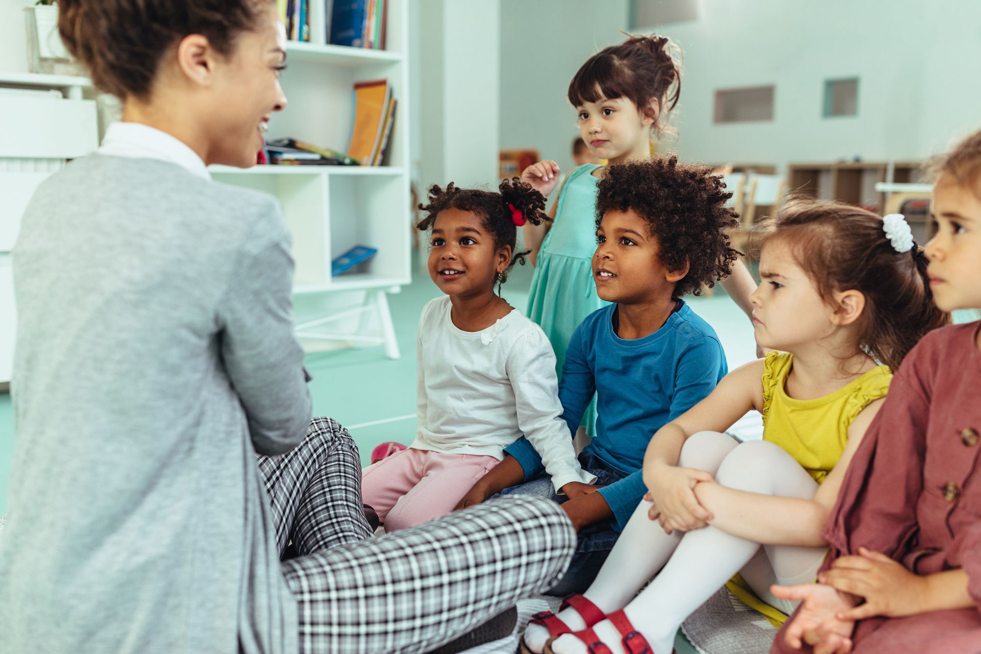 A teacher sits on the floor leading a small group of attentive children in a classroom setting.