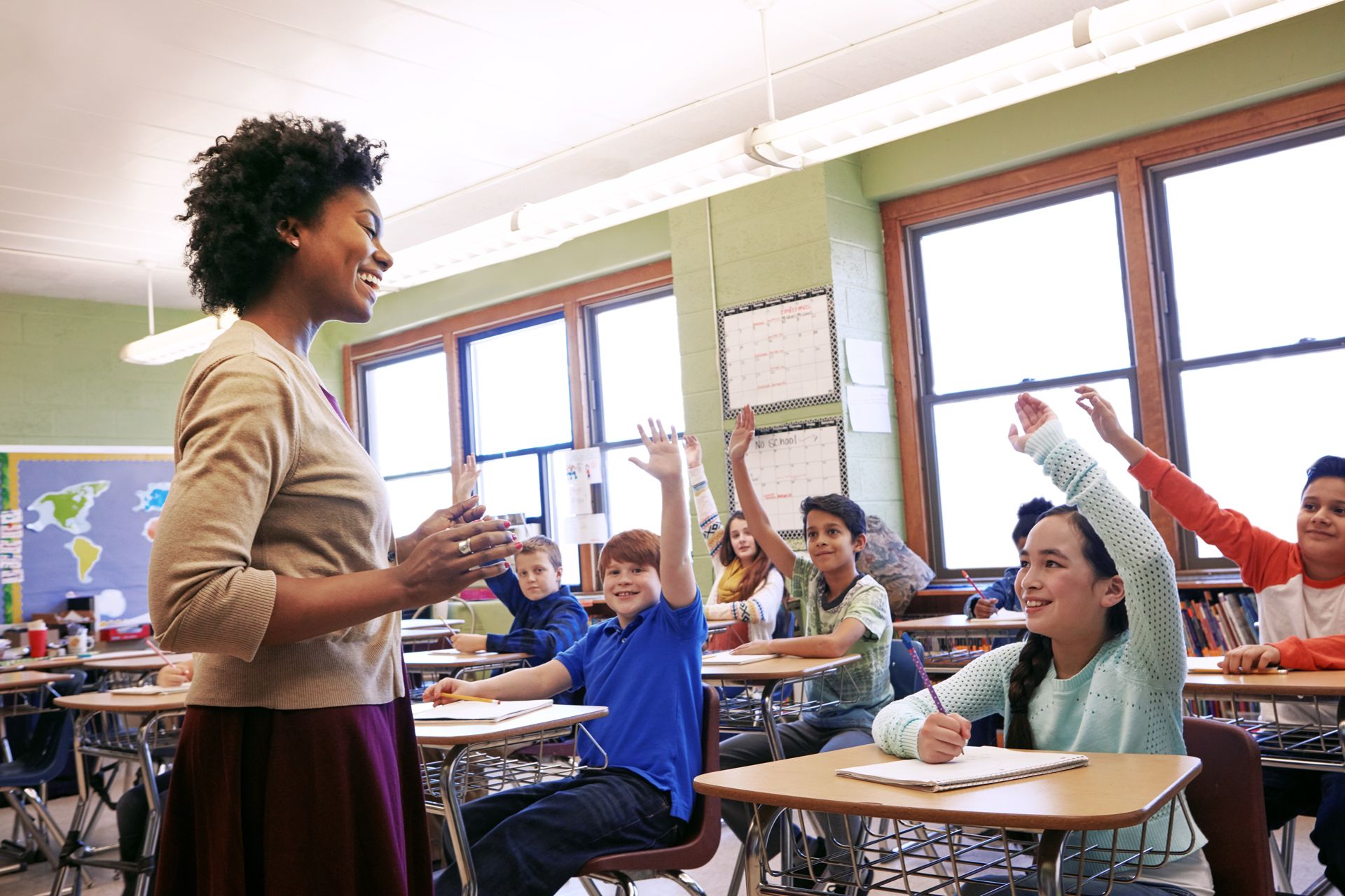 A teacher smiles at students with their hands raised in a sunlit classroom.