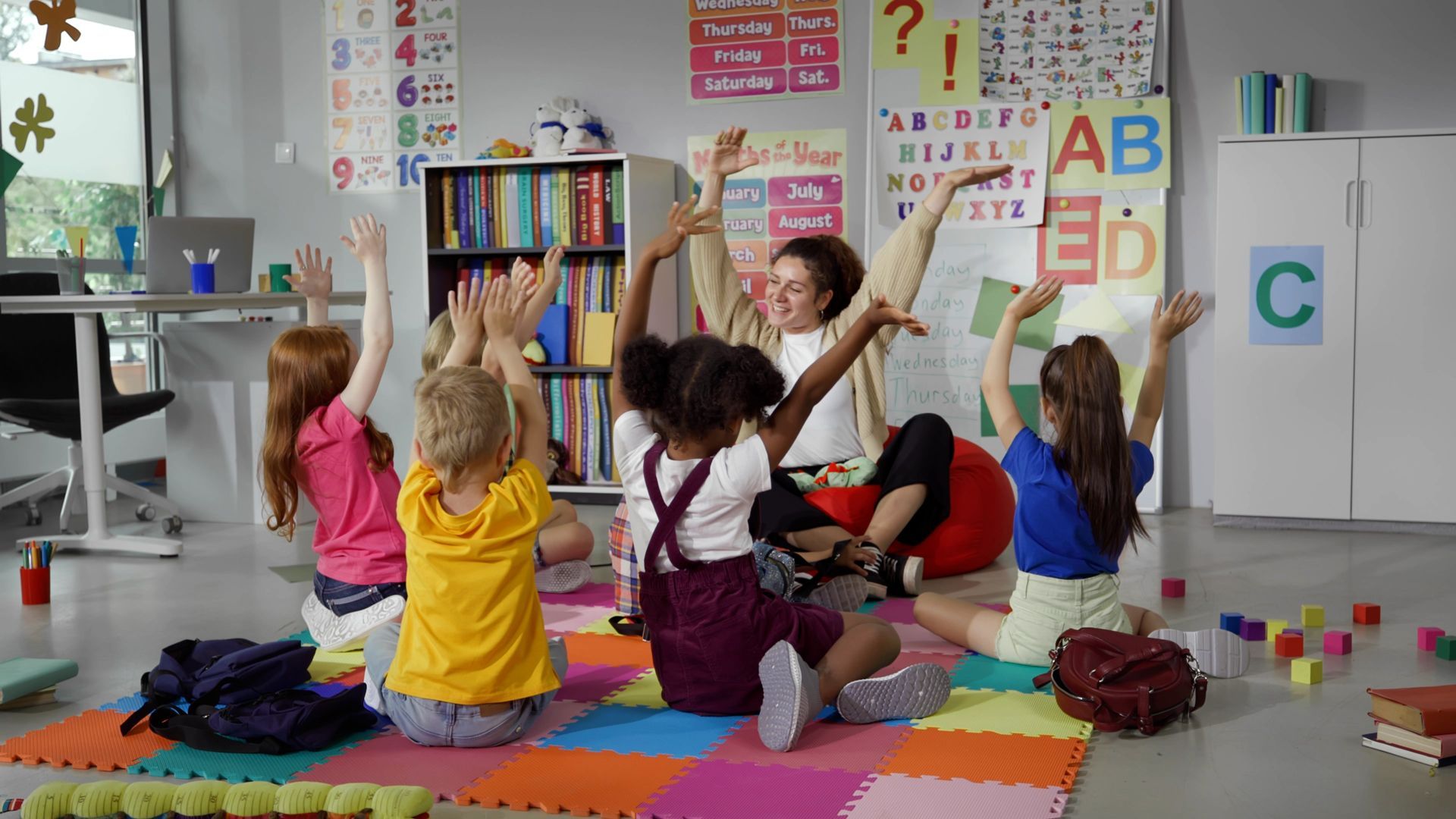 A teacher and four children sit on a colorful mat in a classroom, all raising their hands with joyful expressions.