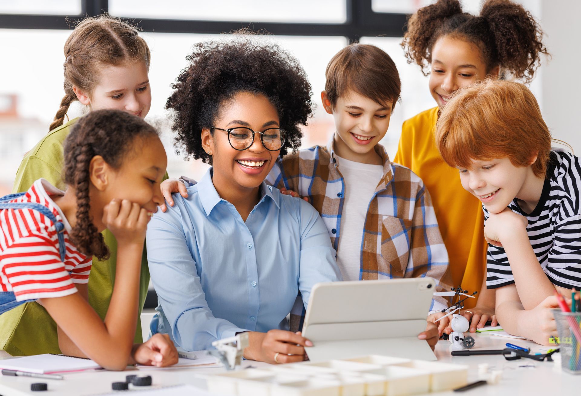 A teacher and five students look at a tablet together in a brightly lit classroom.