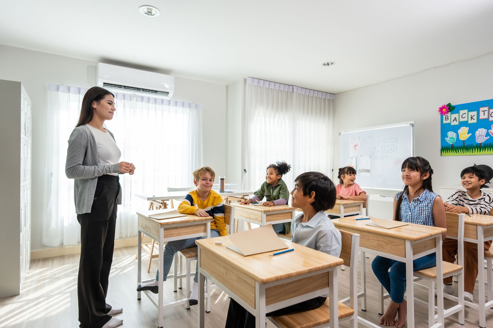 A teacher stands at the front of a brightly lit classroom as a diverse group of students sit at wooden desks.
