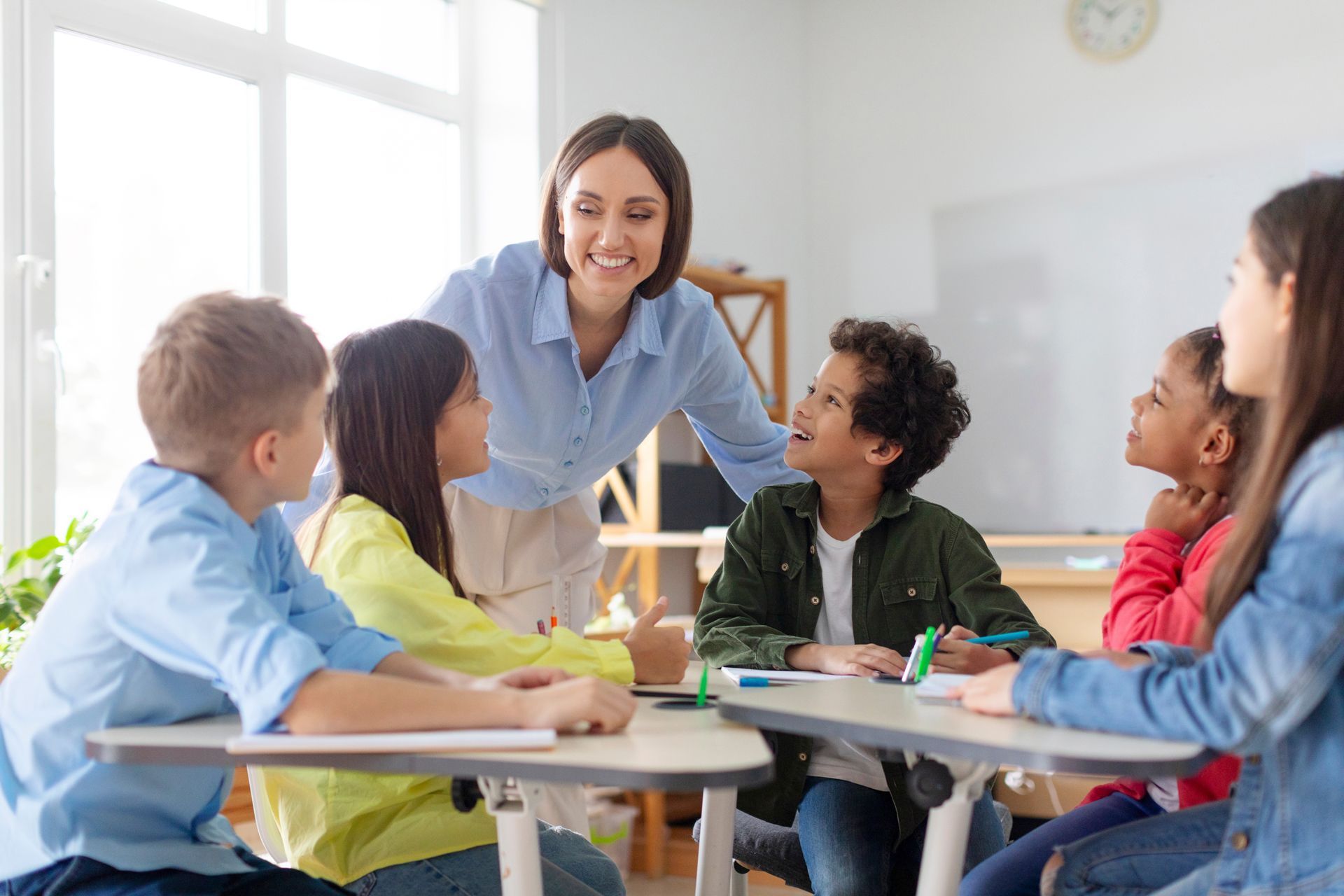A teacher smiles while interacting with a diverse group of students sitting around desks in a brightly lit classroom.